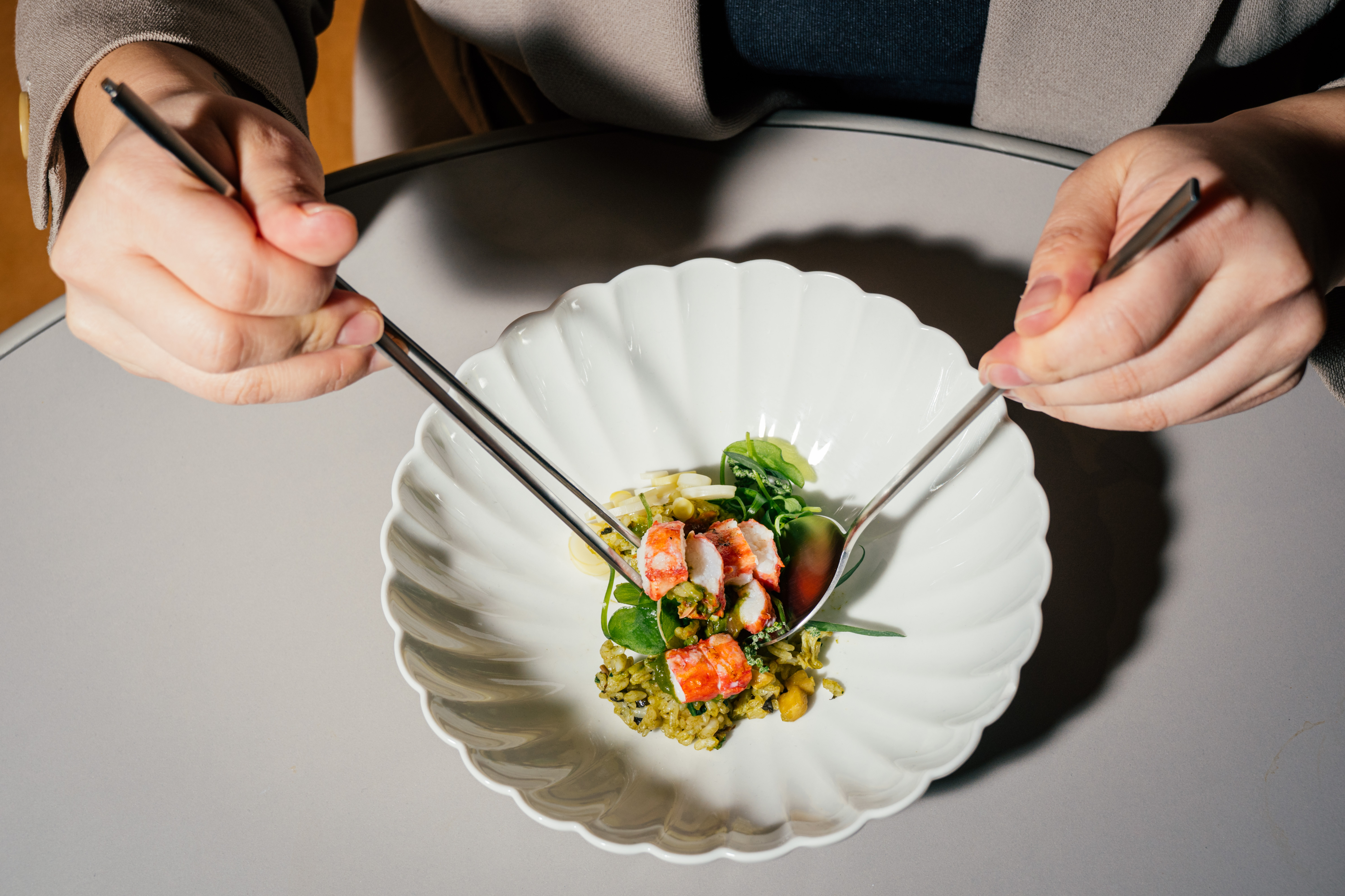 A hand holding a spoon and a pair of chopsticks mixes a bowl of bibimbap.