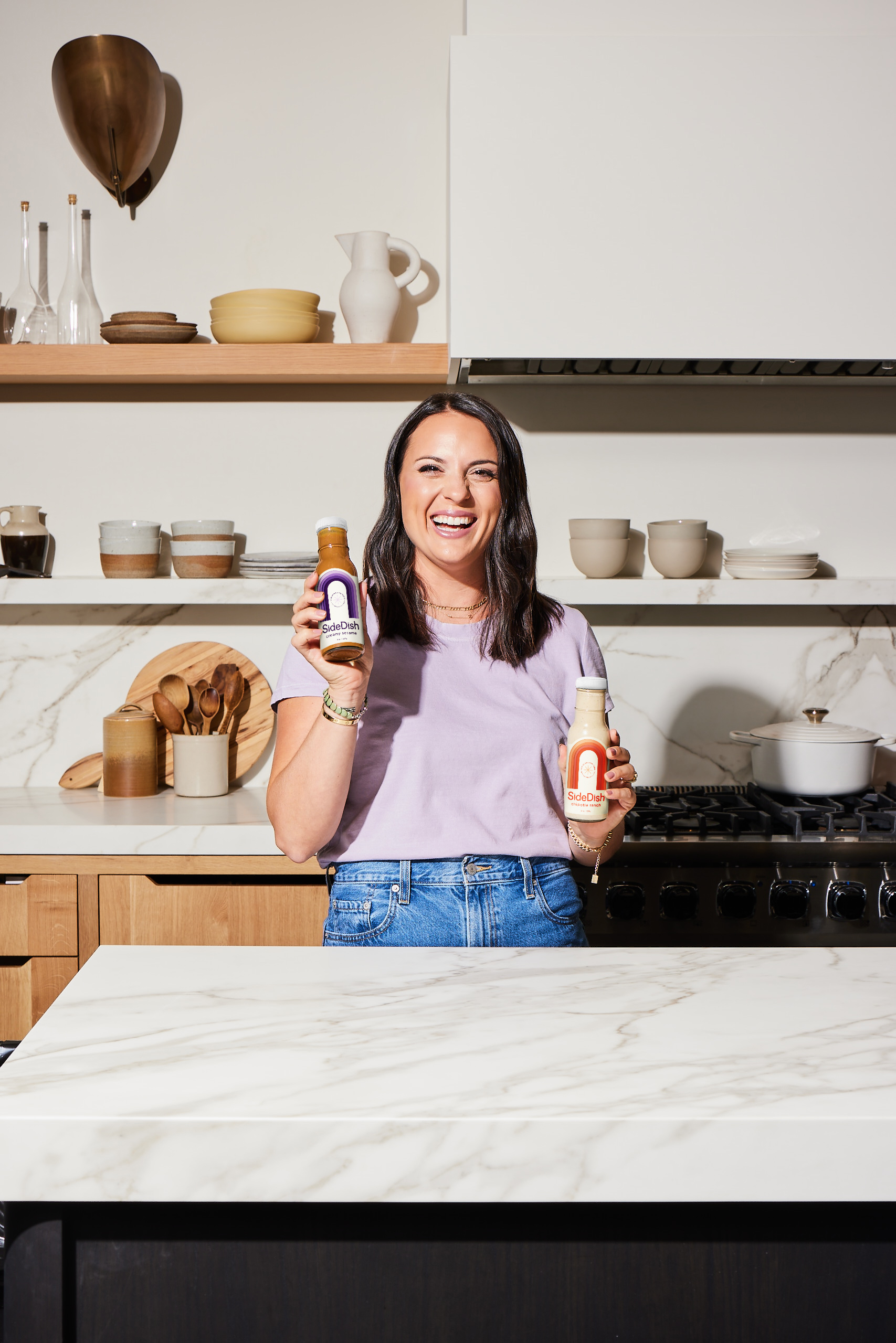 A woman stands in a kitchen, laughing and holding up two bottles of salad dressing.