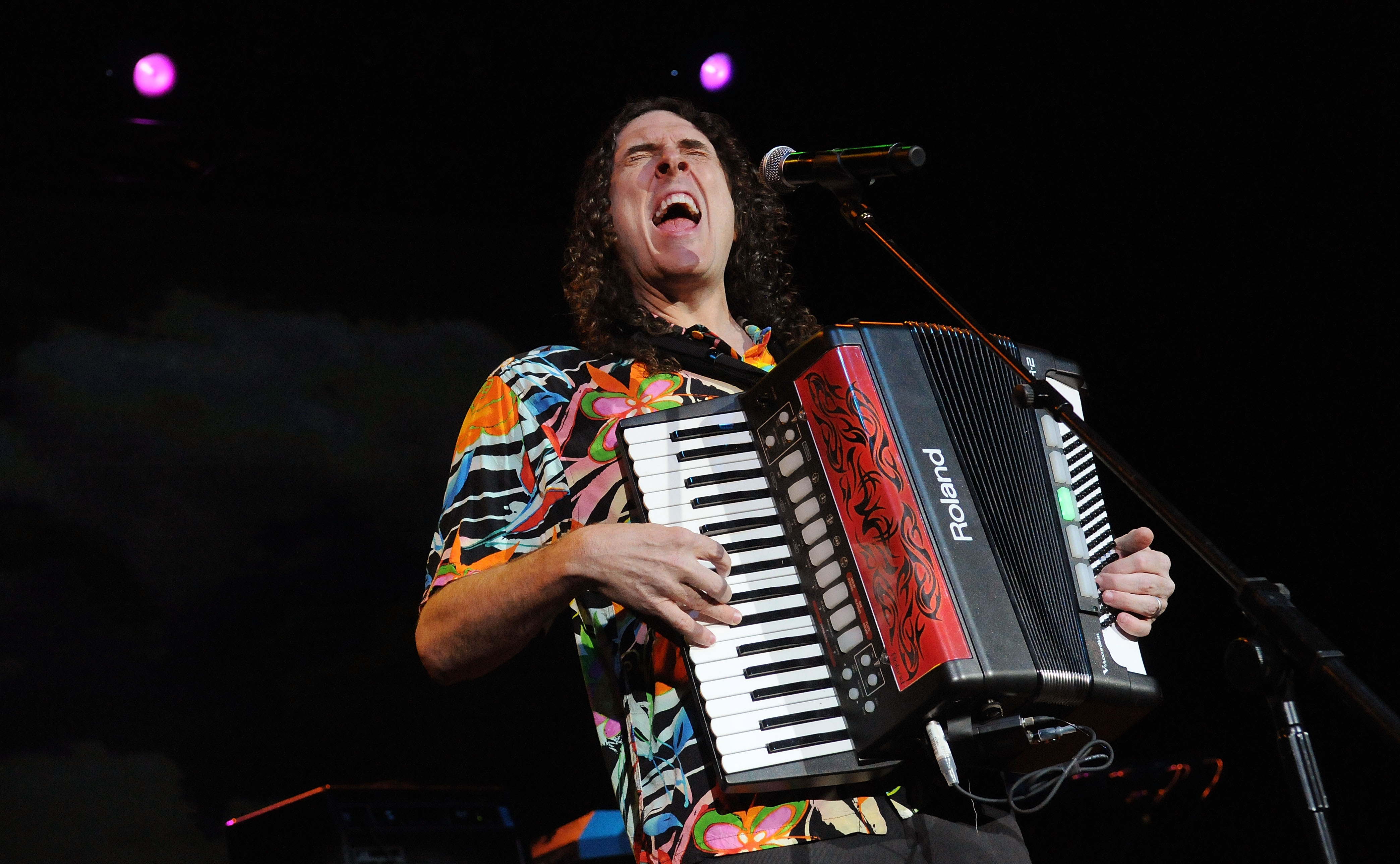 Weird Al Yankovic in a hawaiian shirt wails a song lyric while jamming out on a red and black accordian