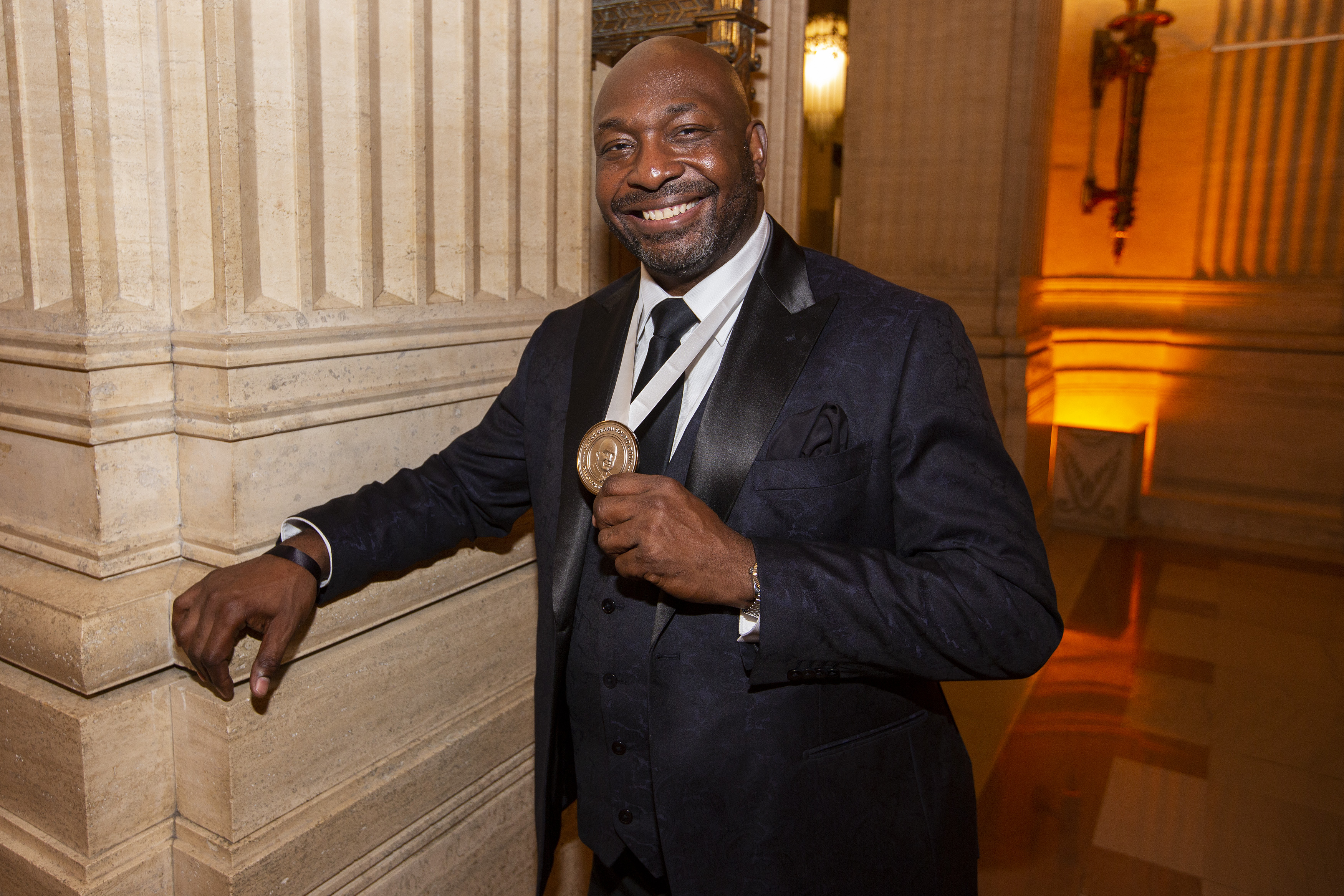 A Black man in a suit poses, smiling, with his James Beard Award medal.