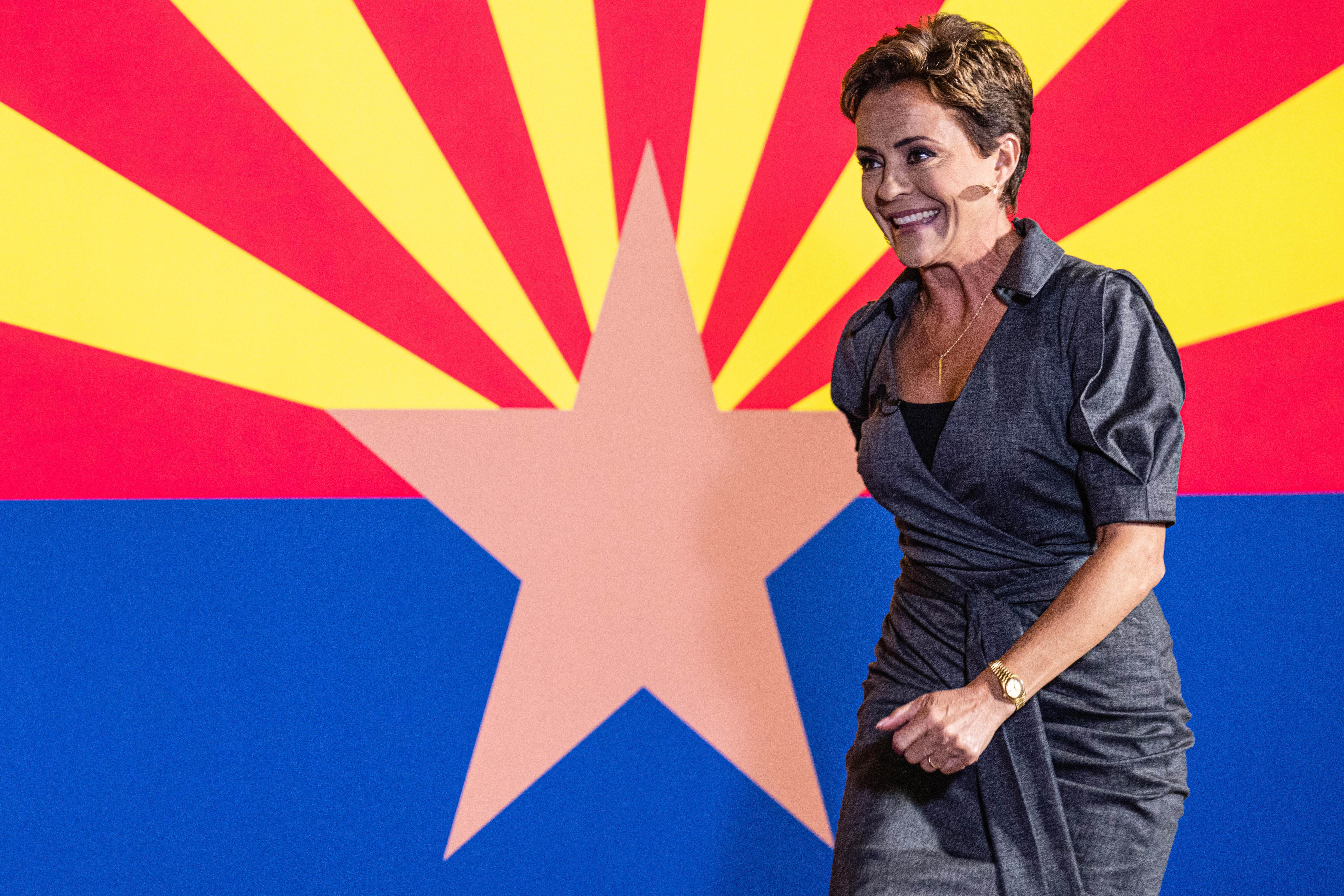 Lake, in a gray wrap dress, her dark blonde hair cut short, smiles as she strides forward purposefully into the spotlight, a giant Arizona state flag hanging behind her.