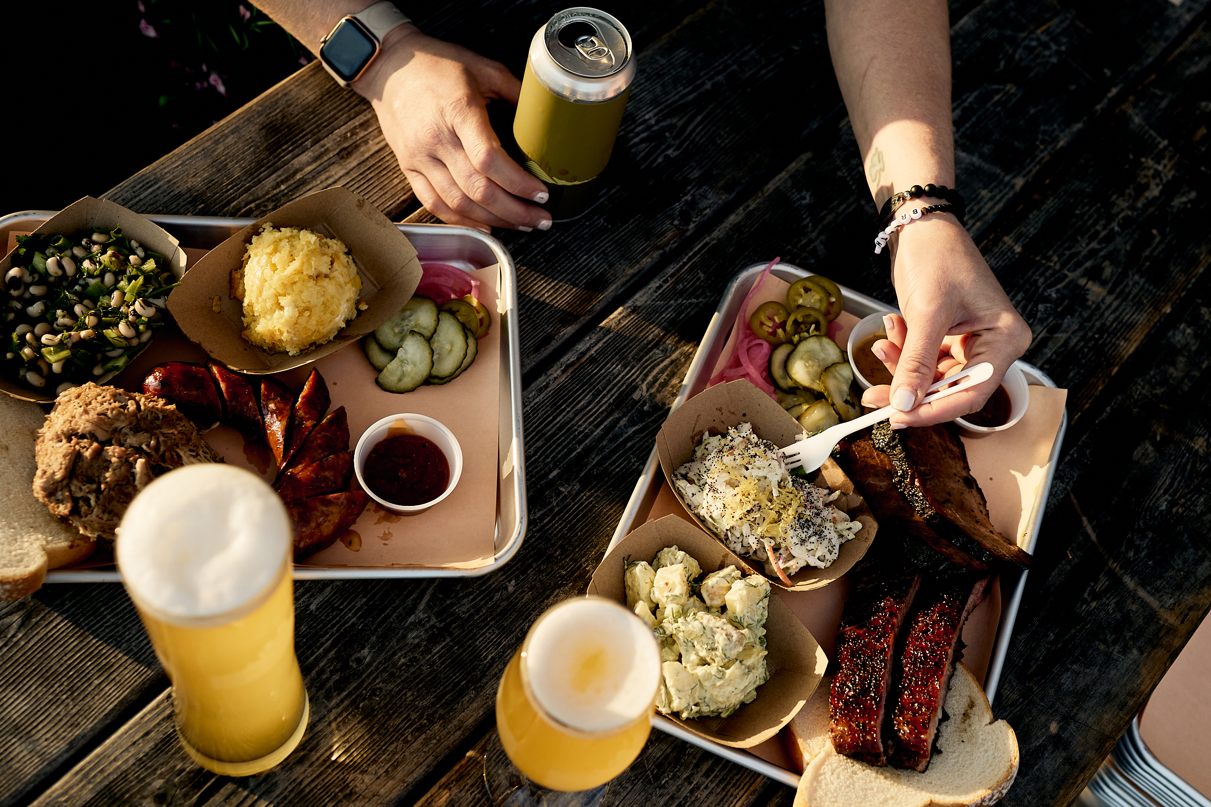 In a top-down view, two trays filled with barbecued meats and side dishes sit on a wooden table next to two glasses and one can of beer; a hand holds the can of beer while another hand reaches for some food with a plastic fork.