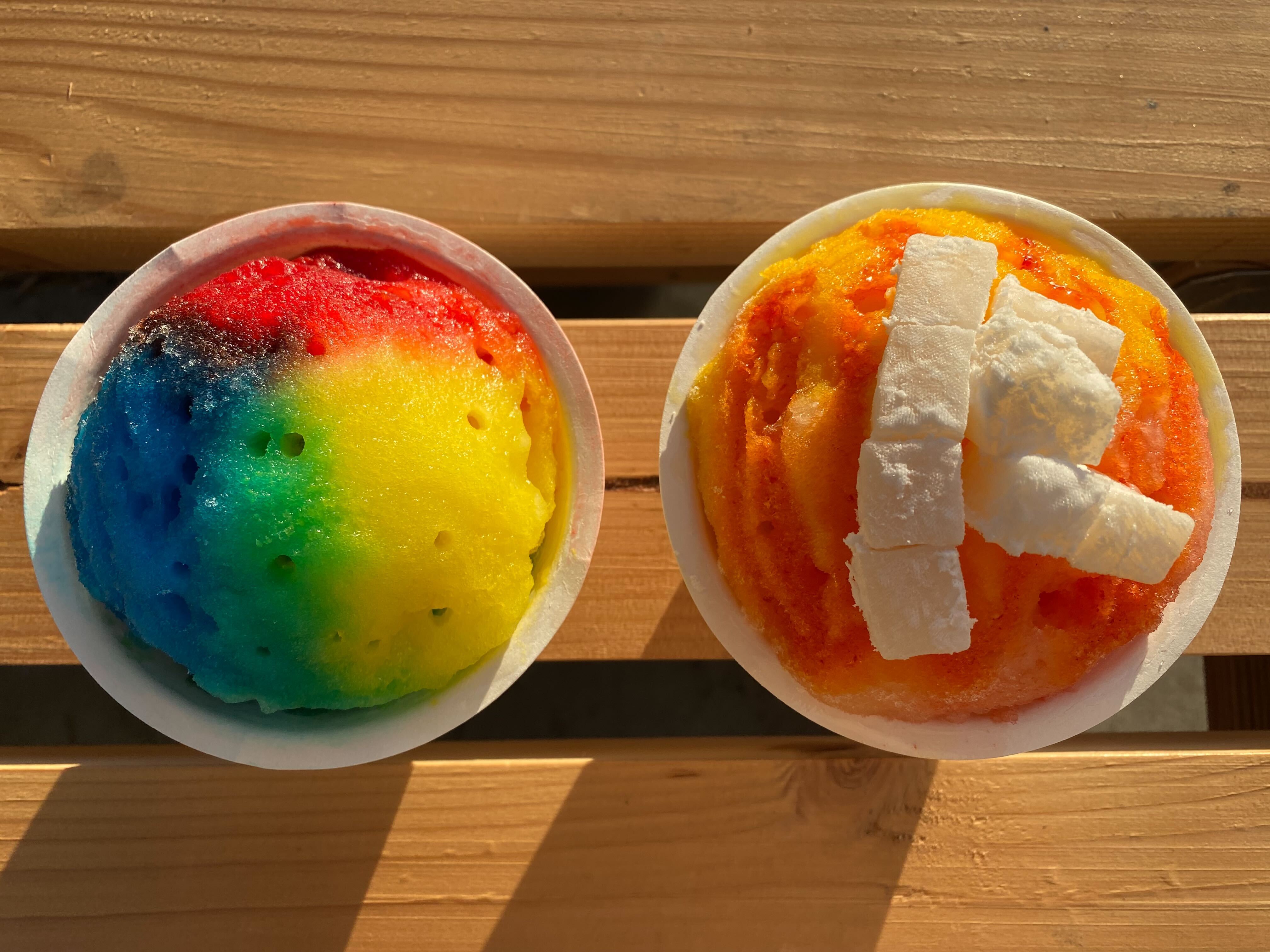 A rainbow covered shave ice cup and an orange-pink shave ice cup with cubes of white mochi on top.