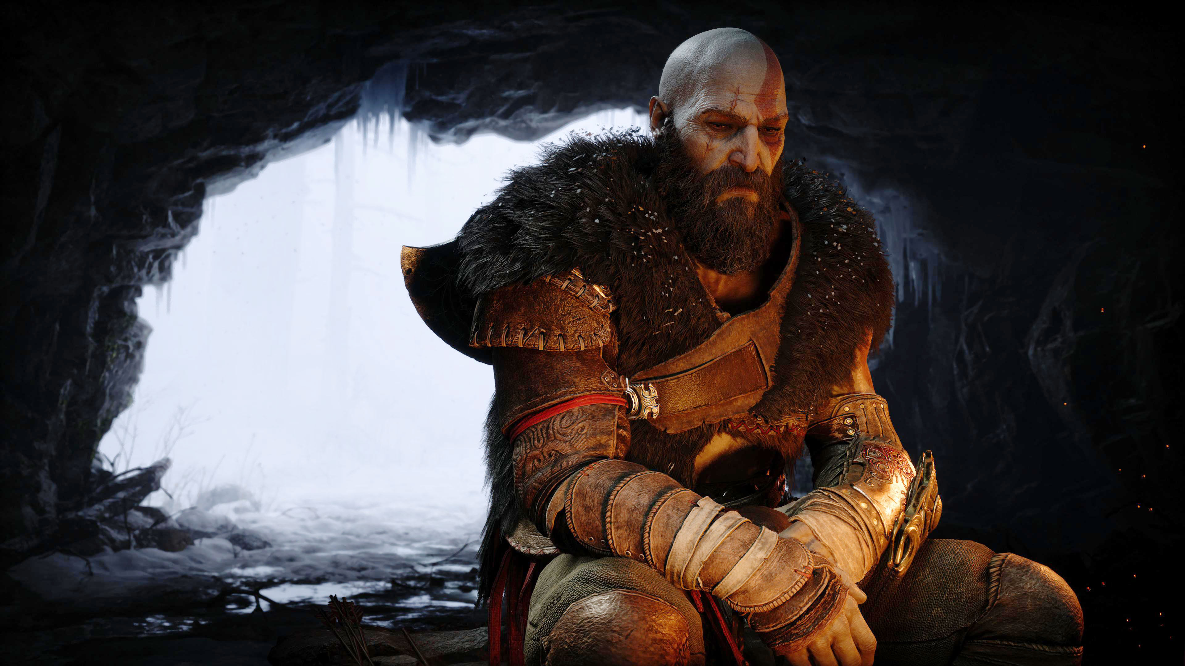Kratos, wearing heavy animal furs and a scowl on his face, sits in front of a fire inside a cave. Behind him, we can see a wintery, cold world in the cave entrance. 