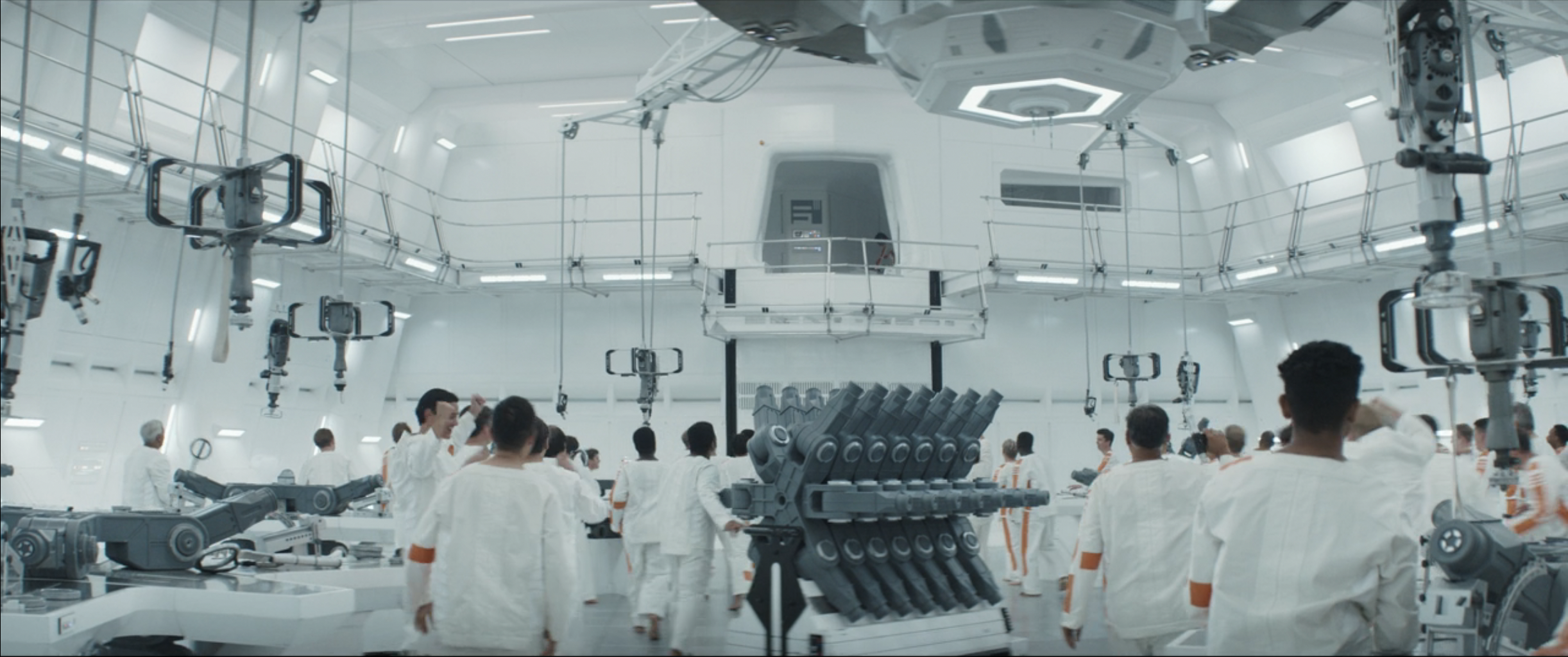 A wide shot of a work floor in Andor’s space prison; prisoners are all looking up at an open door with a rack of mechanical joints in the middle