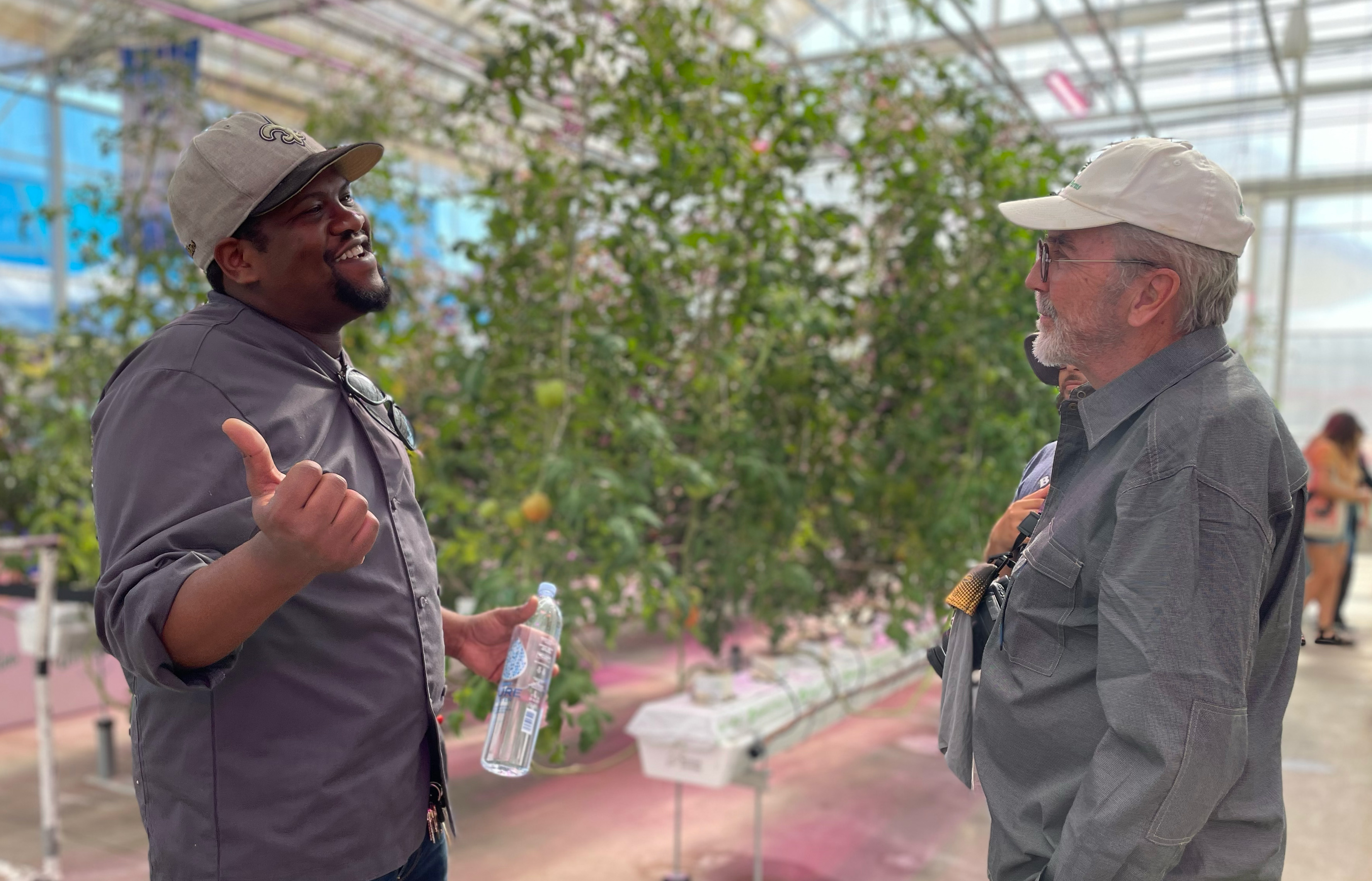 Two men talk in a greenhouse, where numerous vegetable-bearing plants grow around them.