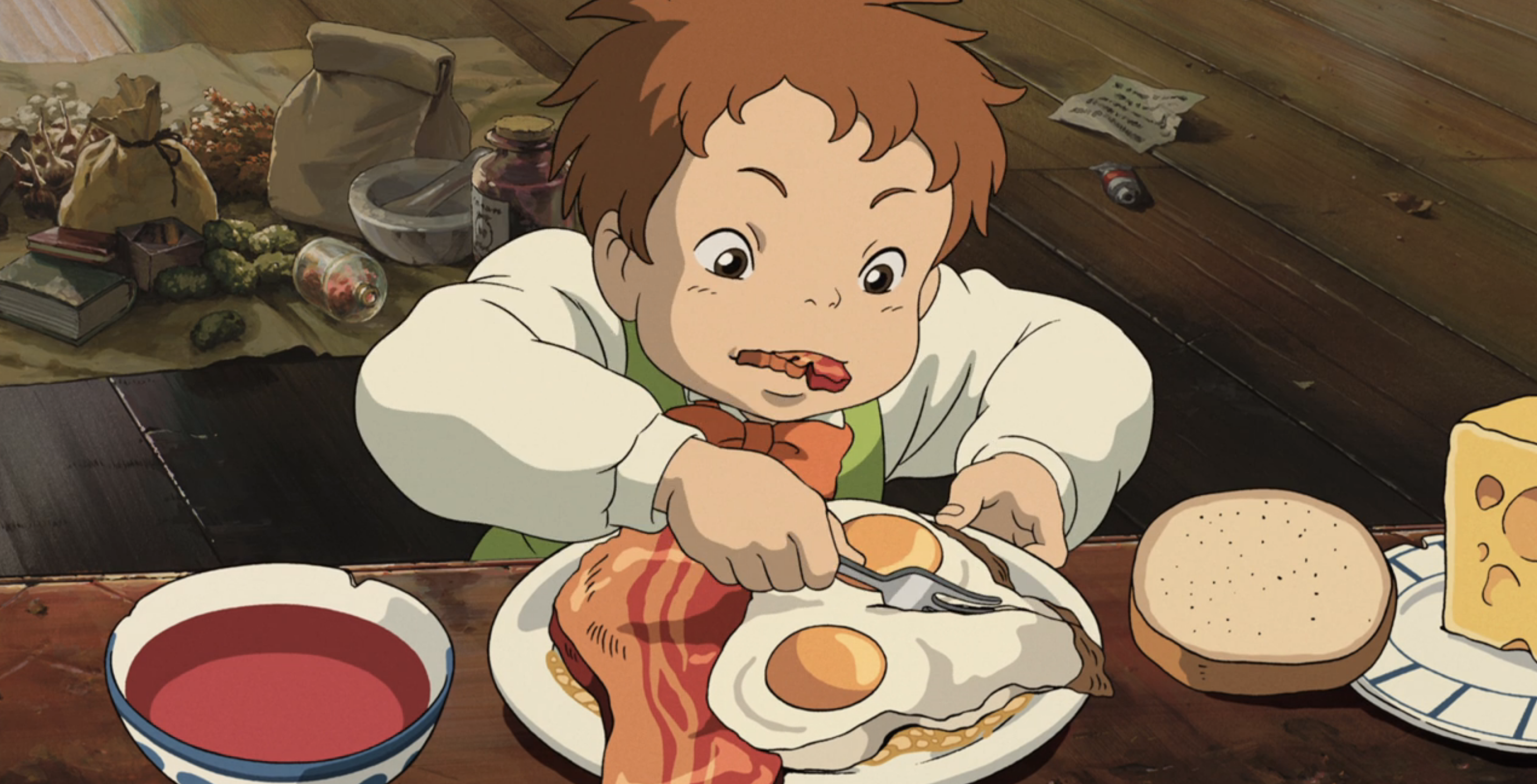 A young red-haired boy chews on a chunk of thick bacon while cutting up his lovingly detailed sunny-side-up eggs with a fork in a scene from Studio Ghibli’s Howl’s Moving Castle
