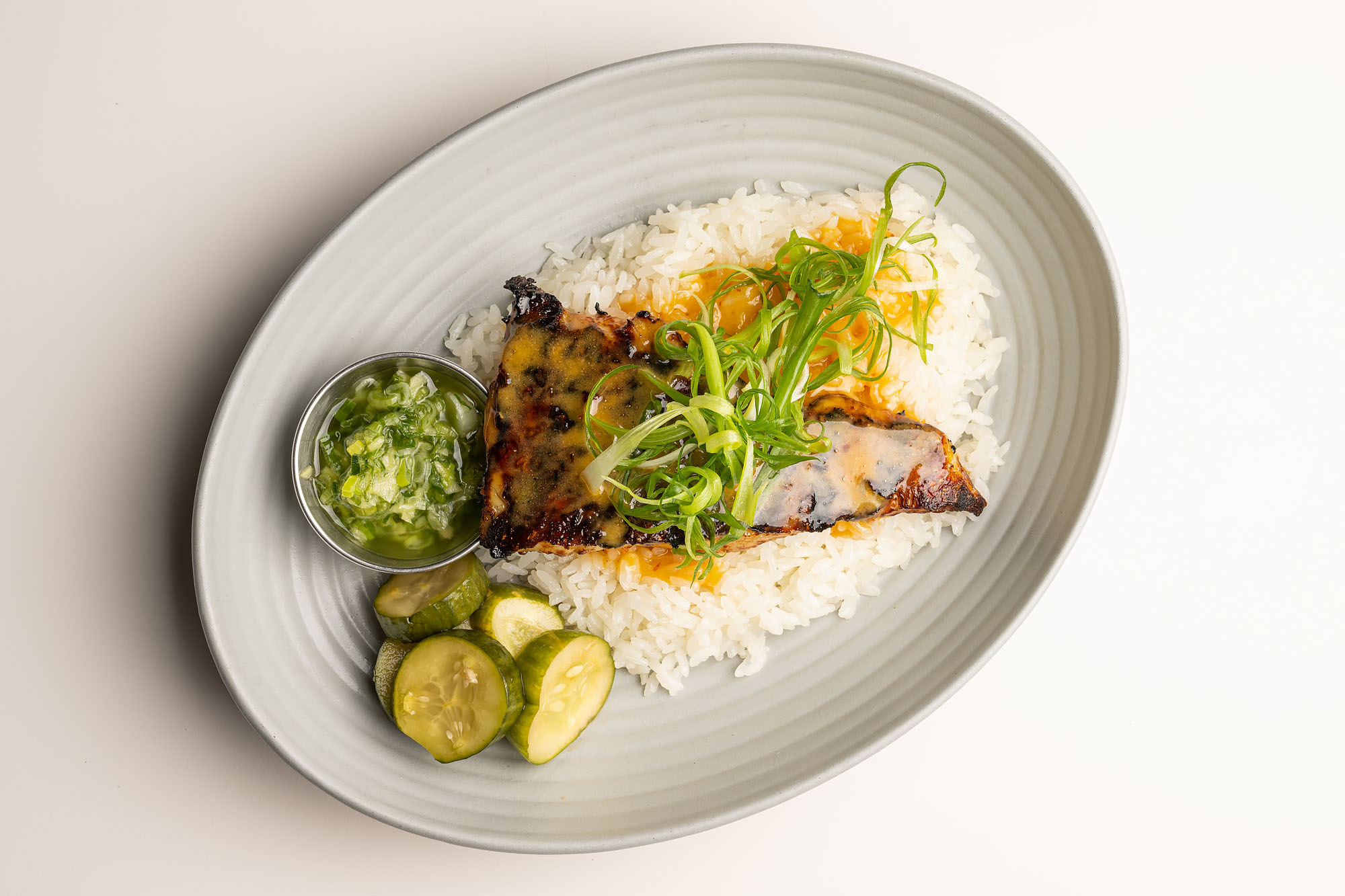 A white plate with white rice, slices of cucumber, a cut of fish covered in an orange sauce, and chopped scallions.