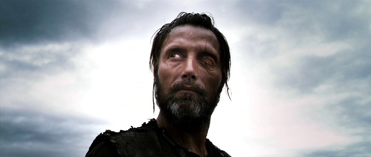 A stern looking man (Mads Mikkelsen) with a missing eyes stares into the distance with a cloudy sky visible behind him.