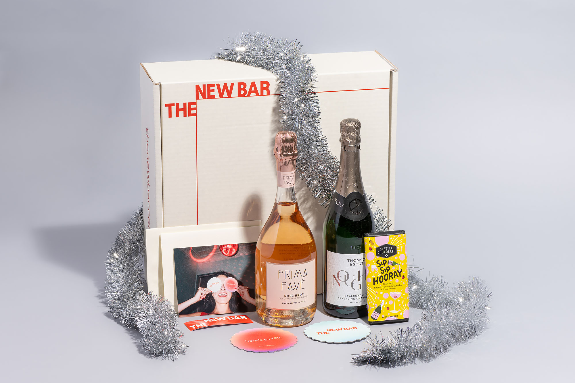 A gift box containing two bottles of sparkling wine and a chocolate bar, wrapped in silver tinsel.