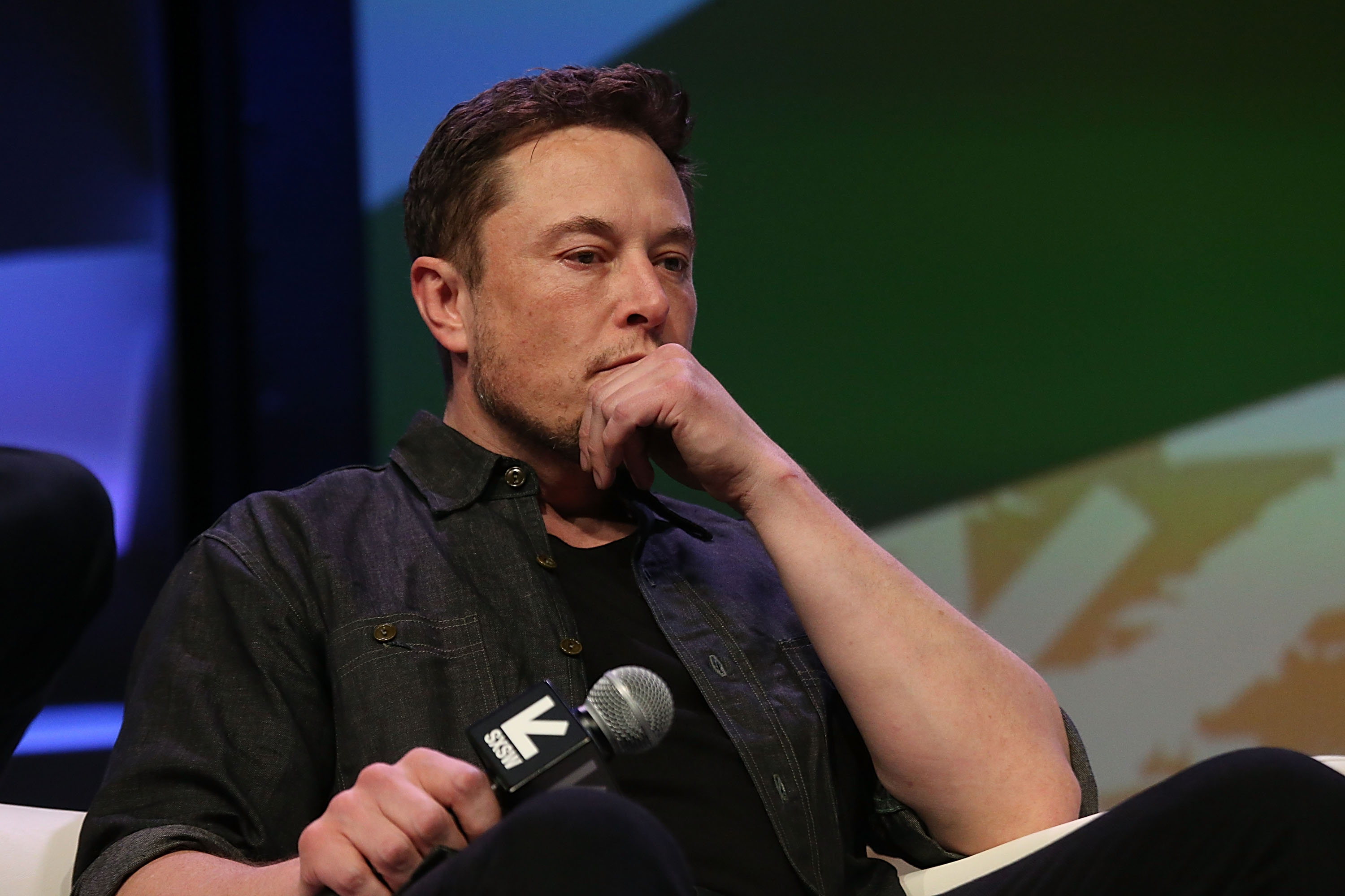 Elon Musk sitting in a chair onstage, holding his chin in one hand and a microphone in the other.