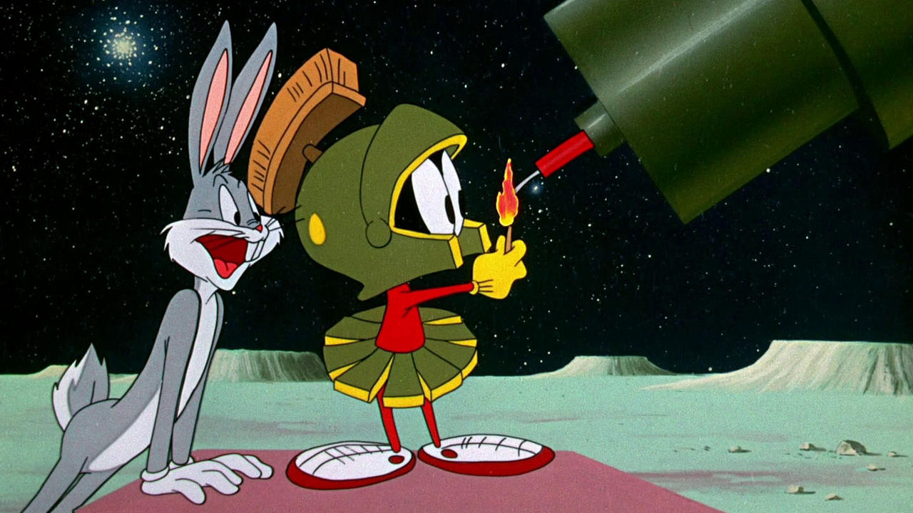 Still frame from a Looney Tunes short; Bugs Bunny pops out of a crater to ask Marvin the Martian, who is lighting the fuse on an explosive device, what’s up.