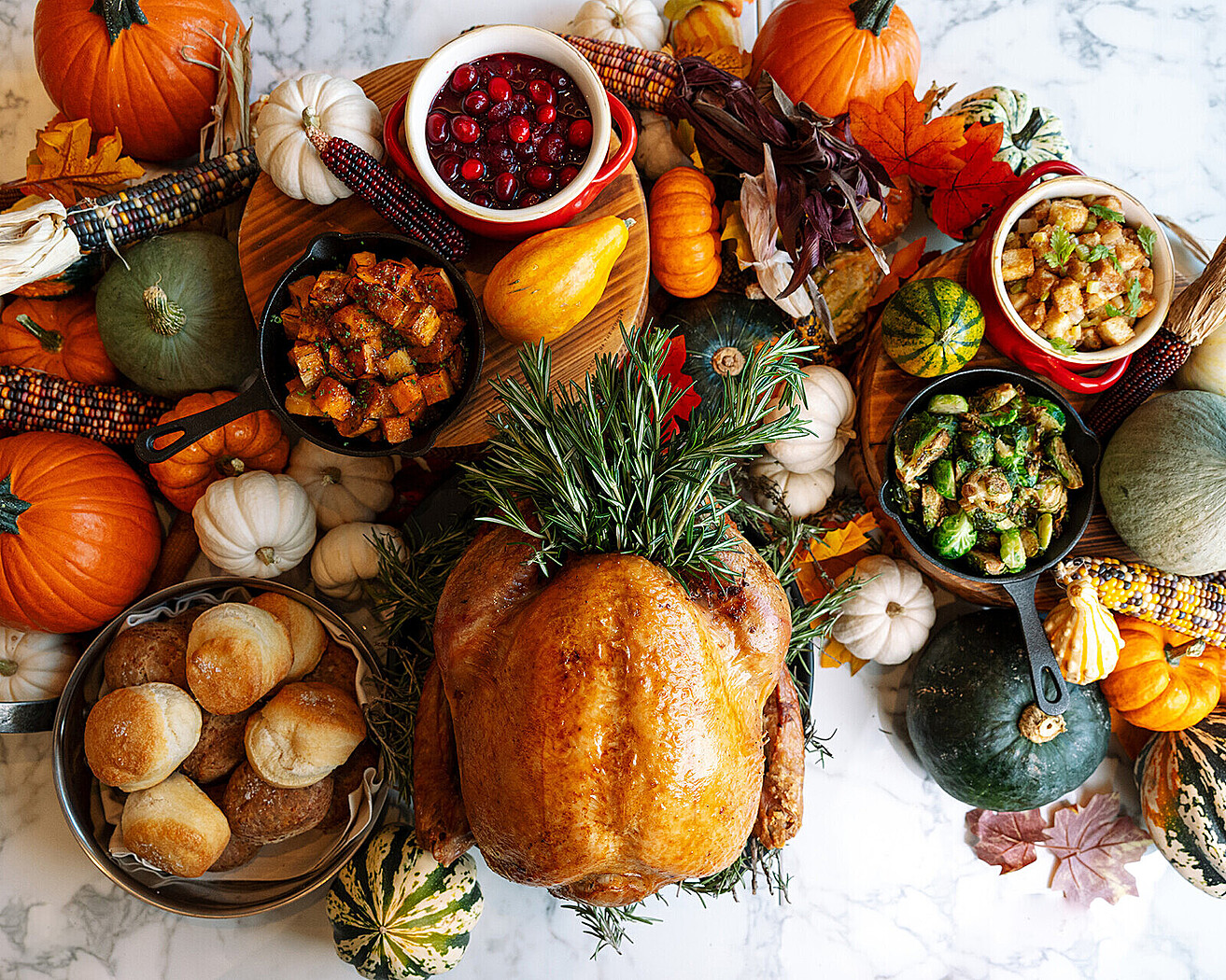 A Thanksgiving turkey table spread with pumpkins, squash, corn, and various dishes filled with vegetables, salads, biscuits, and cranberries