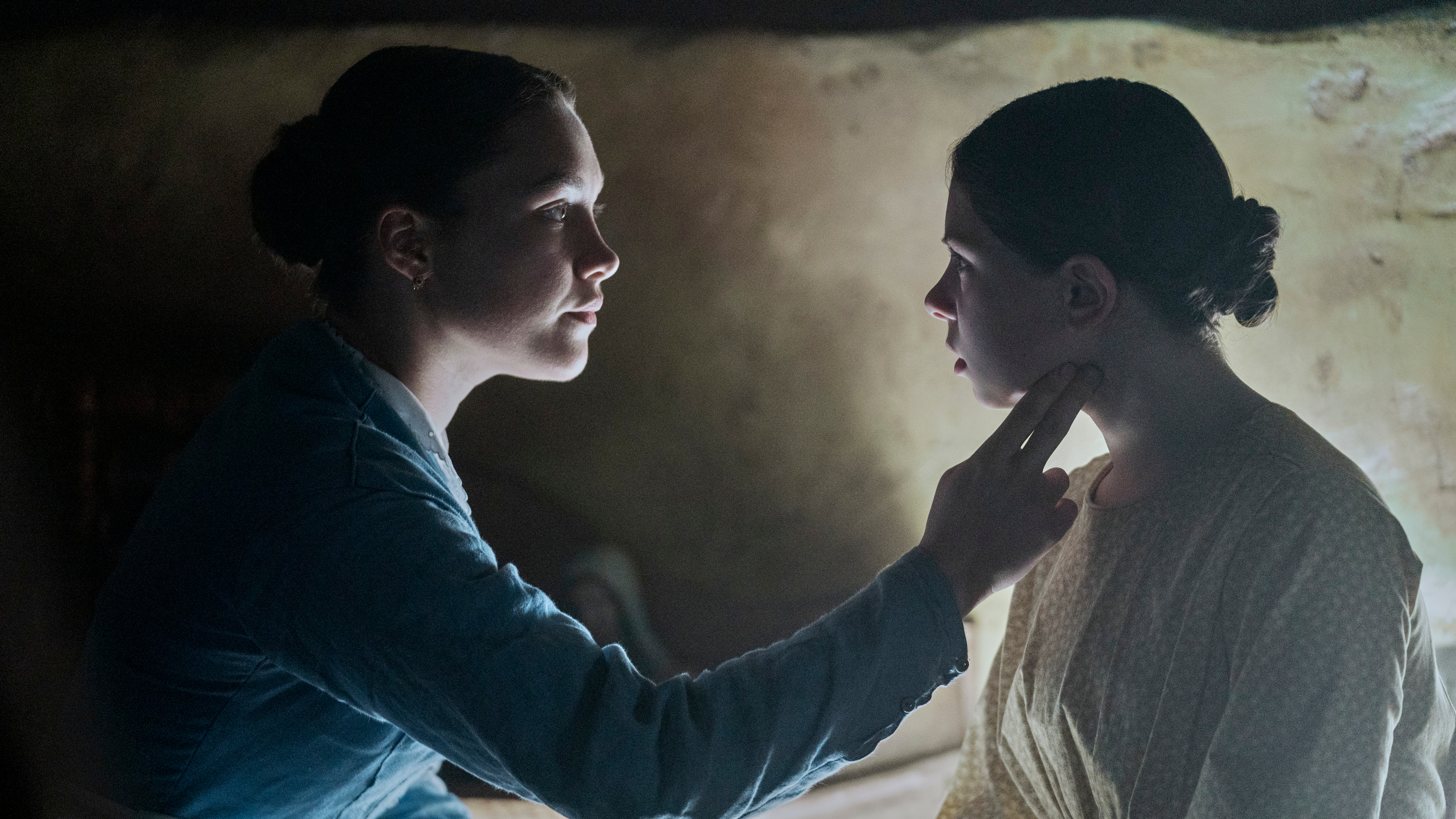 Lib Wright (Florence Pugh) checks the pulse of her patient Anna O’Donnell (Kíla Lord Cassidy) by pressing a finger to her throat in a dark, candlelit room in The Wonder