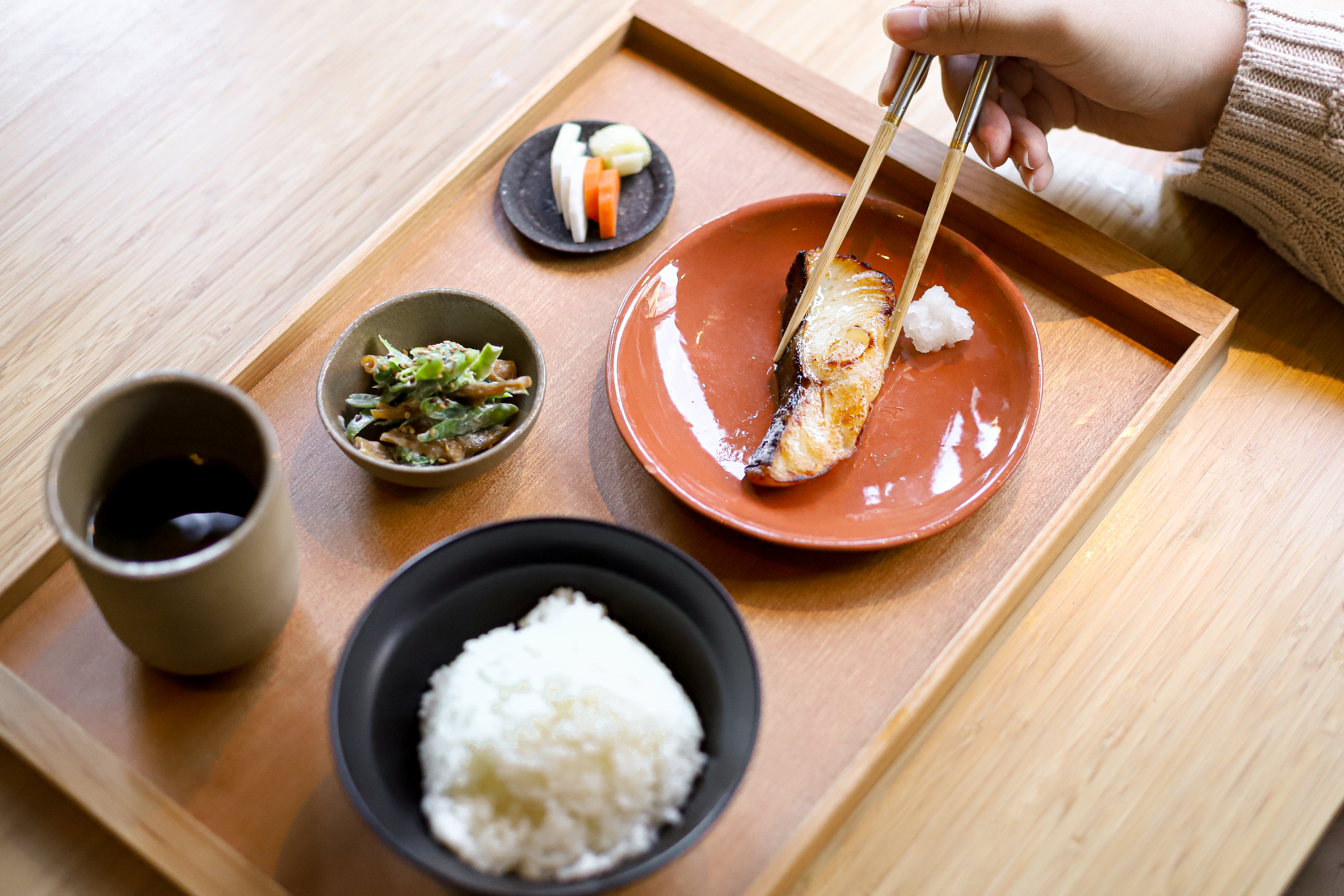 A piece of fish is picked up with a pair of chopsticks from a tray of food.