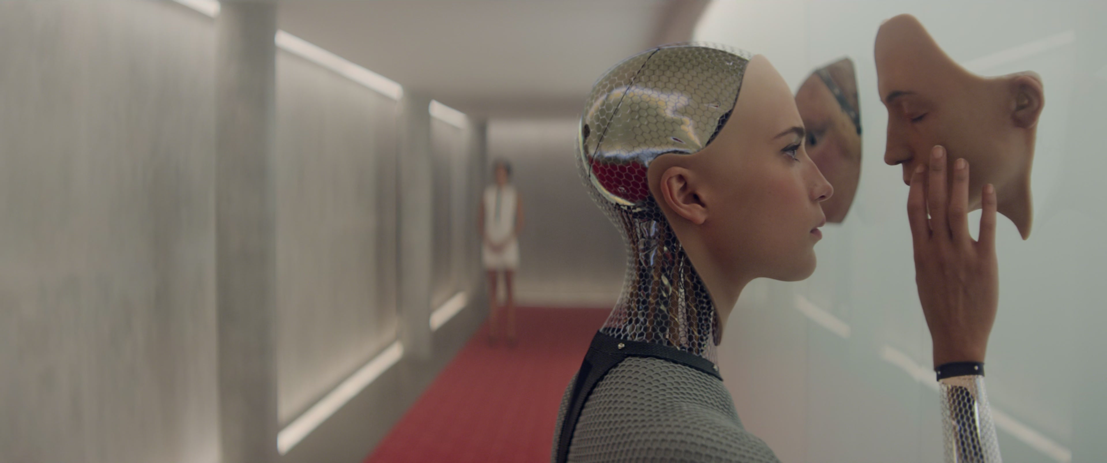 An android woman touching an artificial face hanging from a wall with a woman in a white dress standing at the far end of a futuristic looking hallway with faded red carpeting.