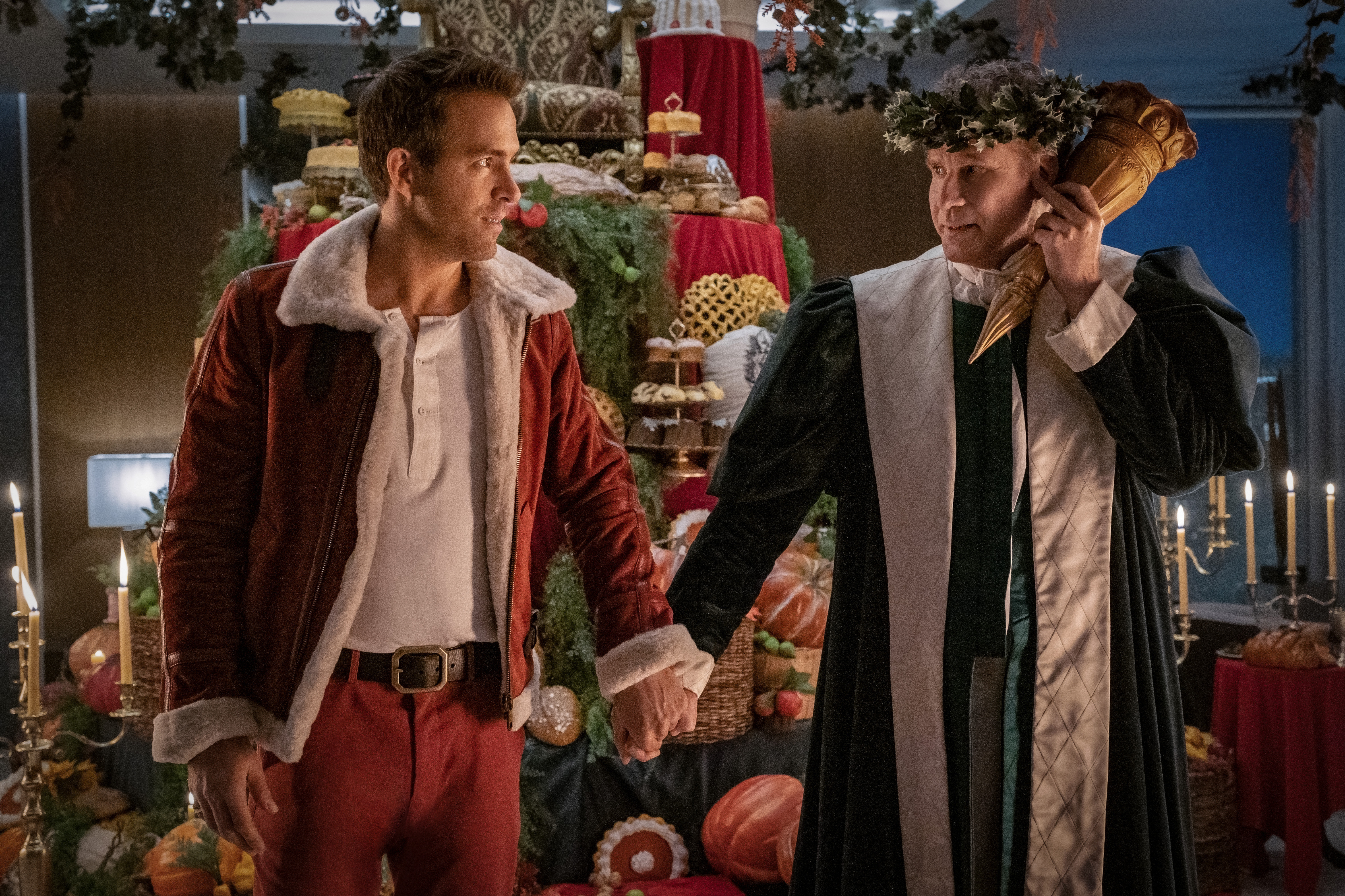 A man wearing a red and white outfit (Ryan Reynolds) holds hands with a man in a green and white outfit (Will Ferrell) with a wreath of holly on his head holding an unlit torch to his ear.