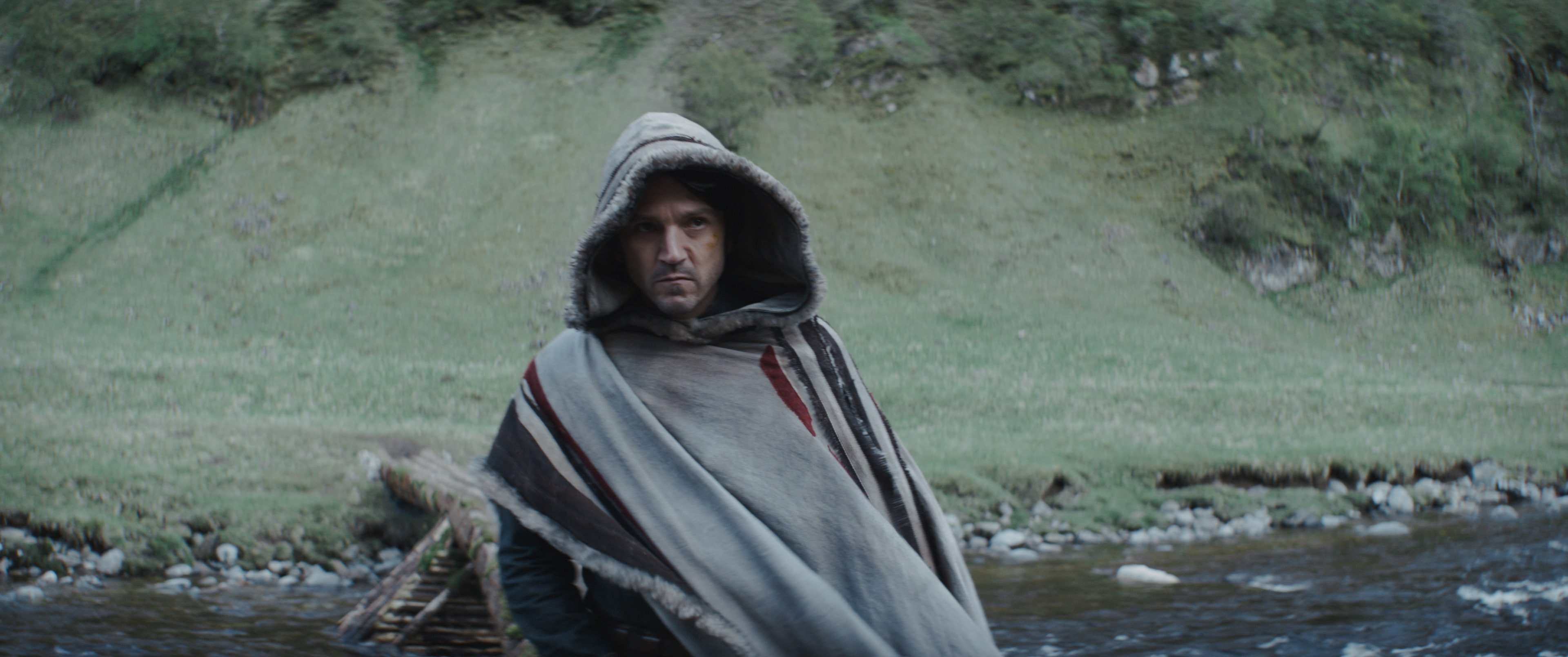 Cassian Andor standing in his poncho glowering at something off-camera. Behind him is a grassy hill