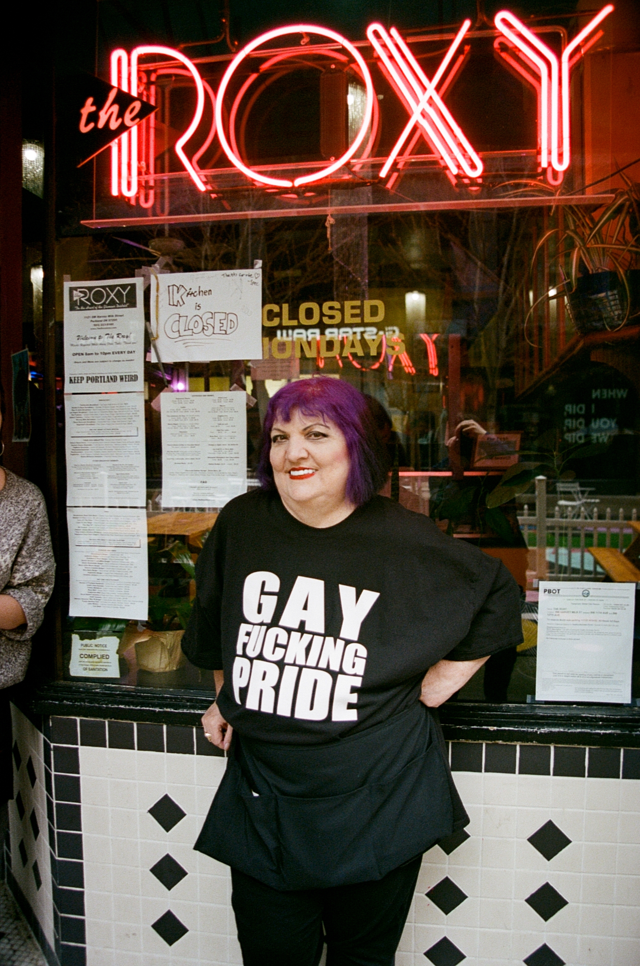 Suzanne Hale wears a black t-shirt that reads “Gay Fucking Pride,” standing outside the Roxy.
