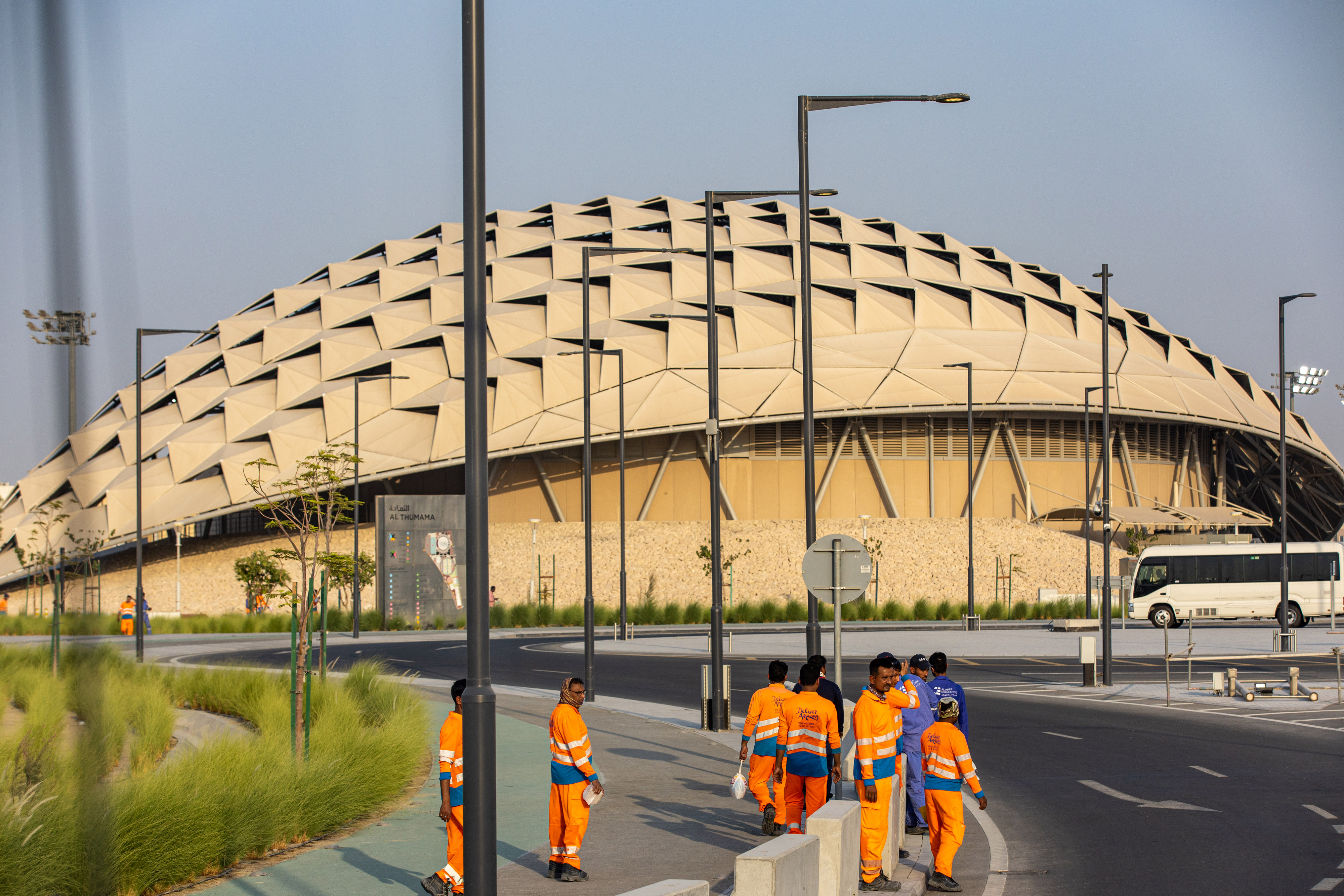 Workers in orange uniforms stand in front of Qatar’s 2022 FIFA World Cup Al Thumama Football Stadium, which is shaped like a cream-colored dome.