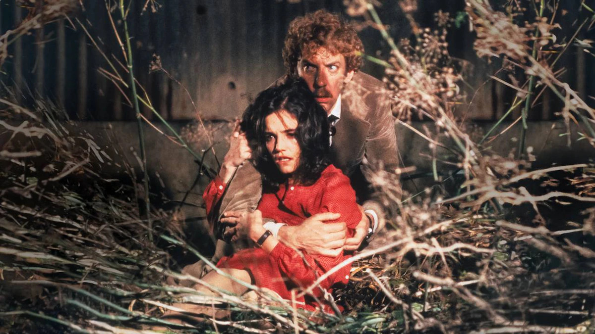 A man in a brown coat (Donald Sutherland) holds a terrified woman in a red dress (Brooke Adams) as they both stare at something concealed by a swath of dead plants and branches.