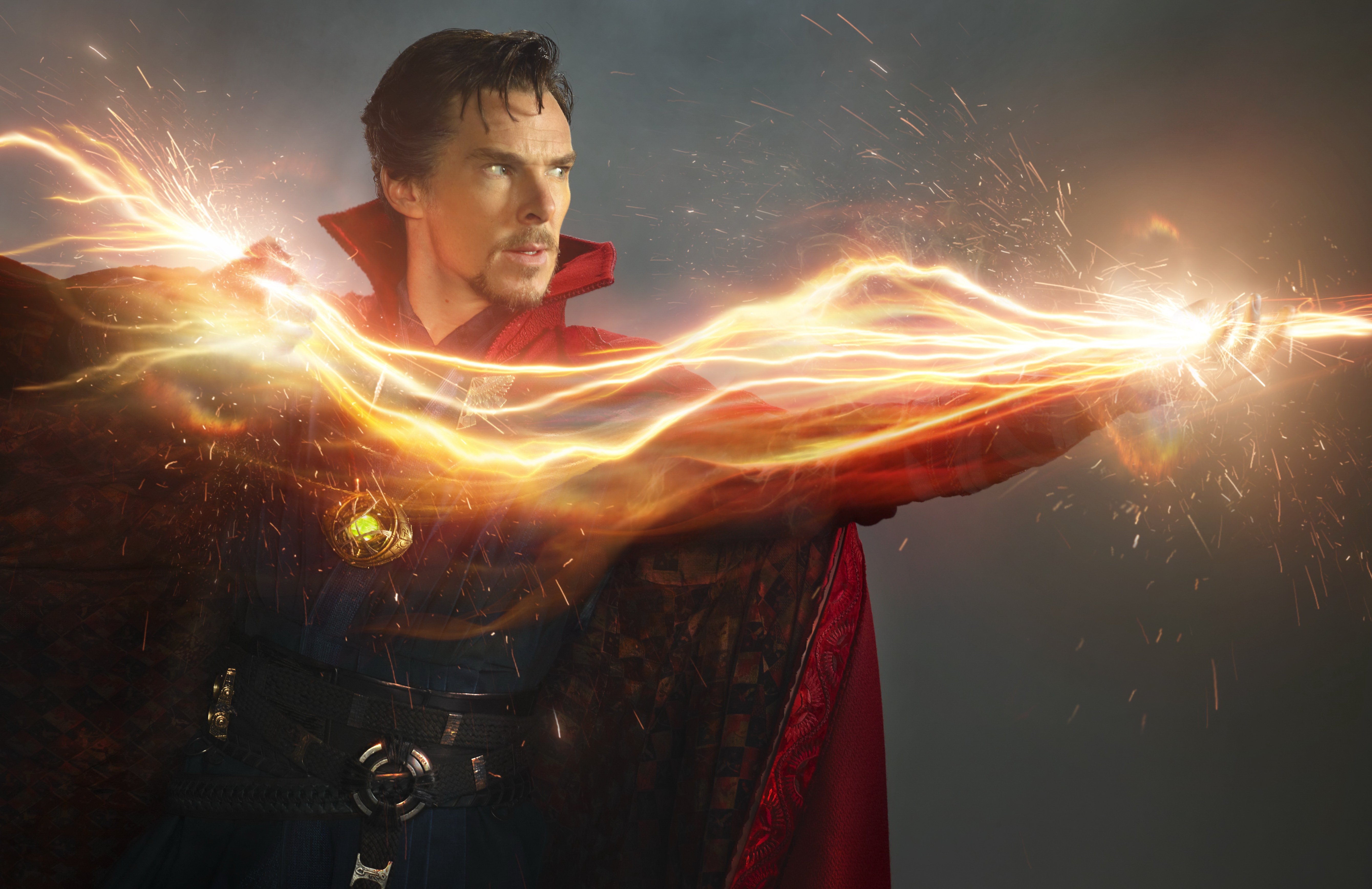 Benedict Cumberbatch as Doctor Strange, in a publicity photo showing him in front of a neutral background, zapping something offscreen with a huge wave of bright orange magical energy