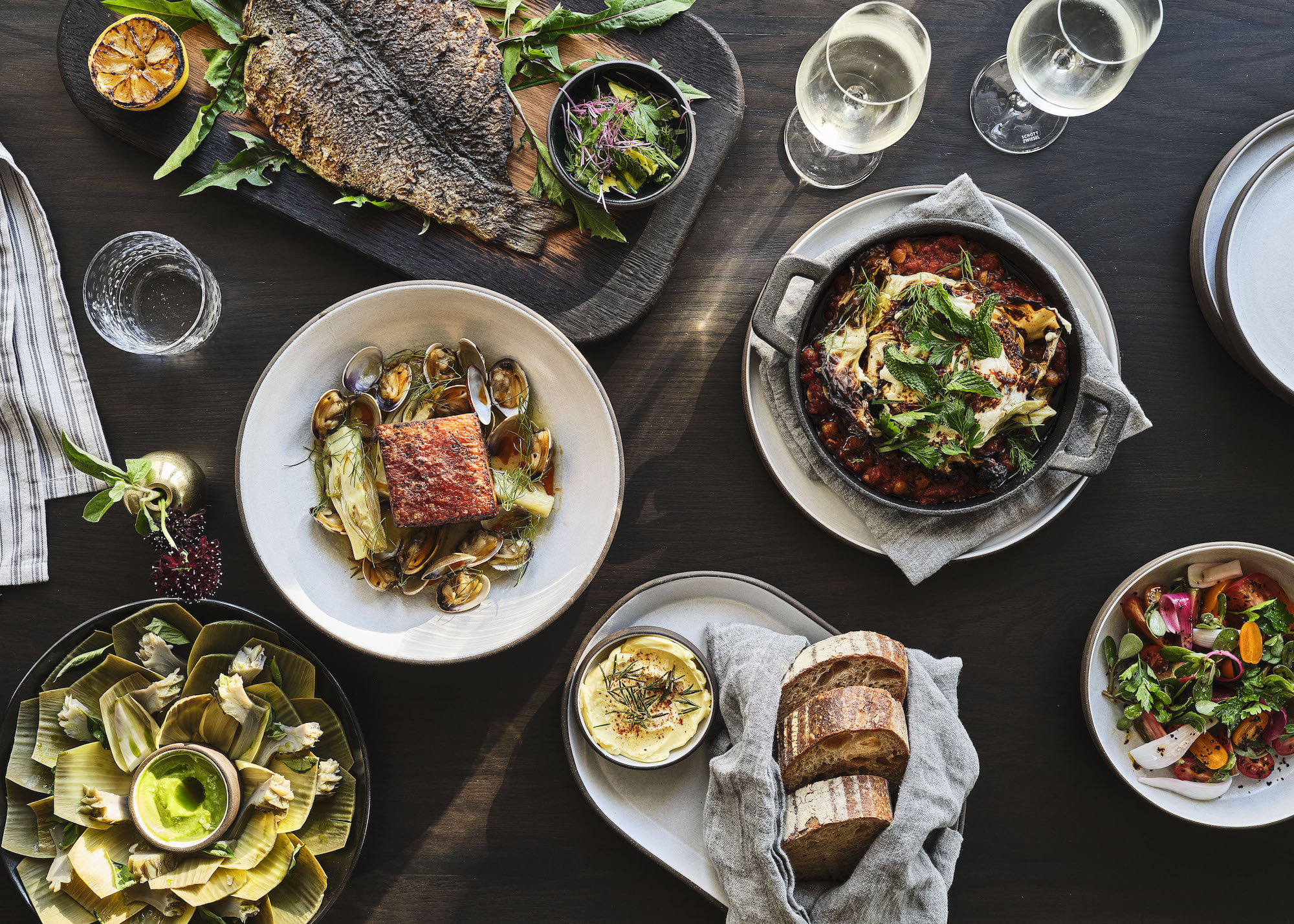 An overhead shot of a crowded dinner table with bread, proteins, and more.