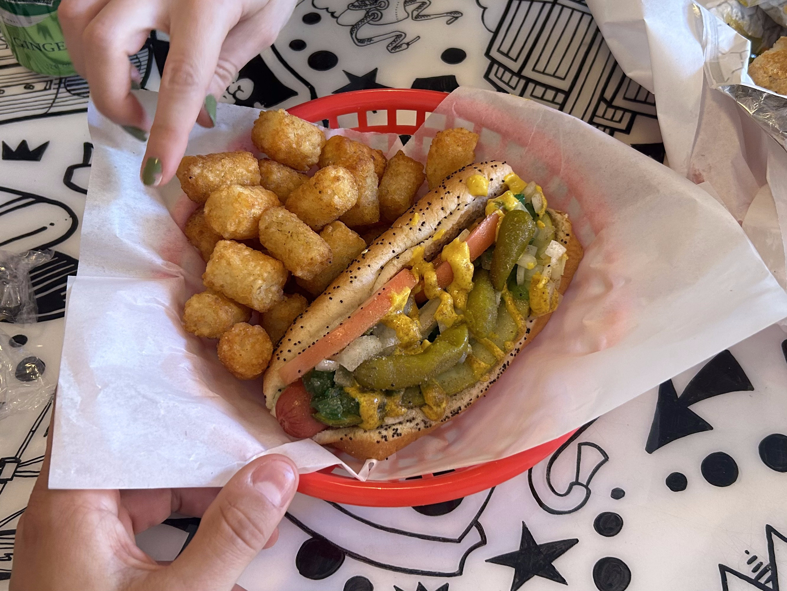 A Chicago-style hot dog with a side of tater tots is served in a red basket with parchment paper.