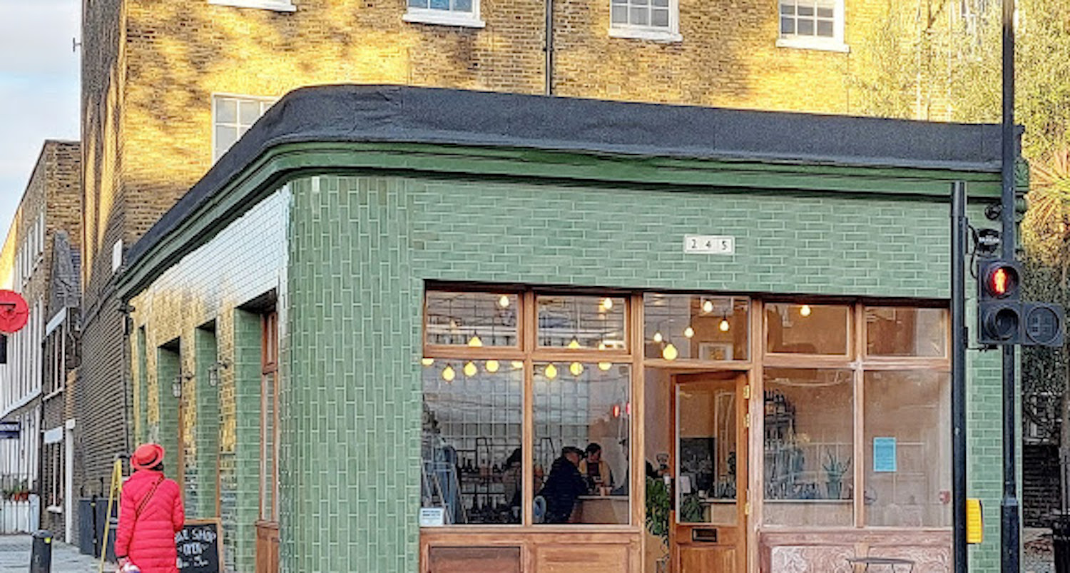 A green-tiled corner unit with wooden framed windows, and a gently curved exterior. A chalkboard sits outside, with a woman in a red coat walking and admiring the frontage.