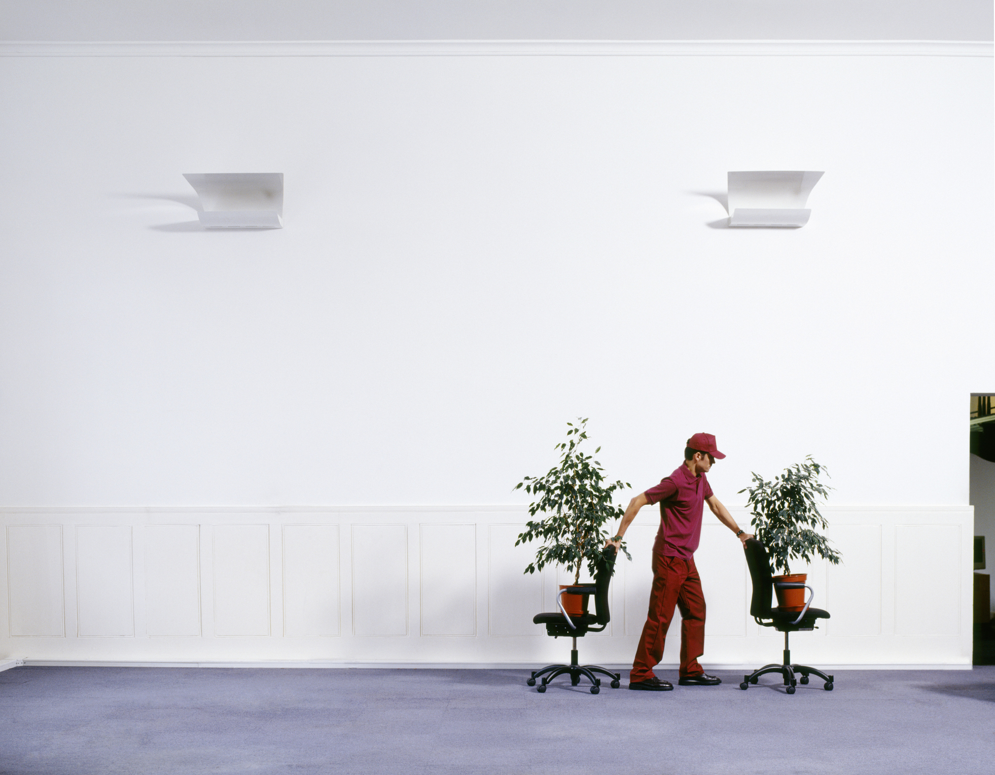 A photo of a man in a worker’s uniform wheeling office chairs, on top of which plants perch, through an empty-looking office setting.