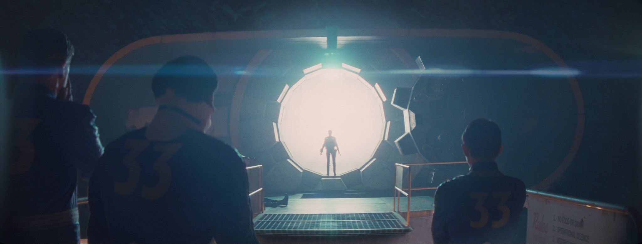 A silhouette stands in front of the blinding white light flooding into an underground shelter from the open vault door of Vault 33 while three people in denim jumpsuits with 33 on the back look on.
