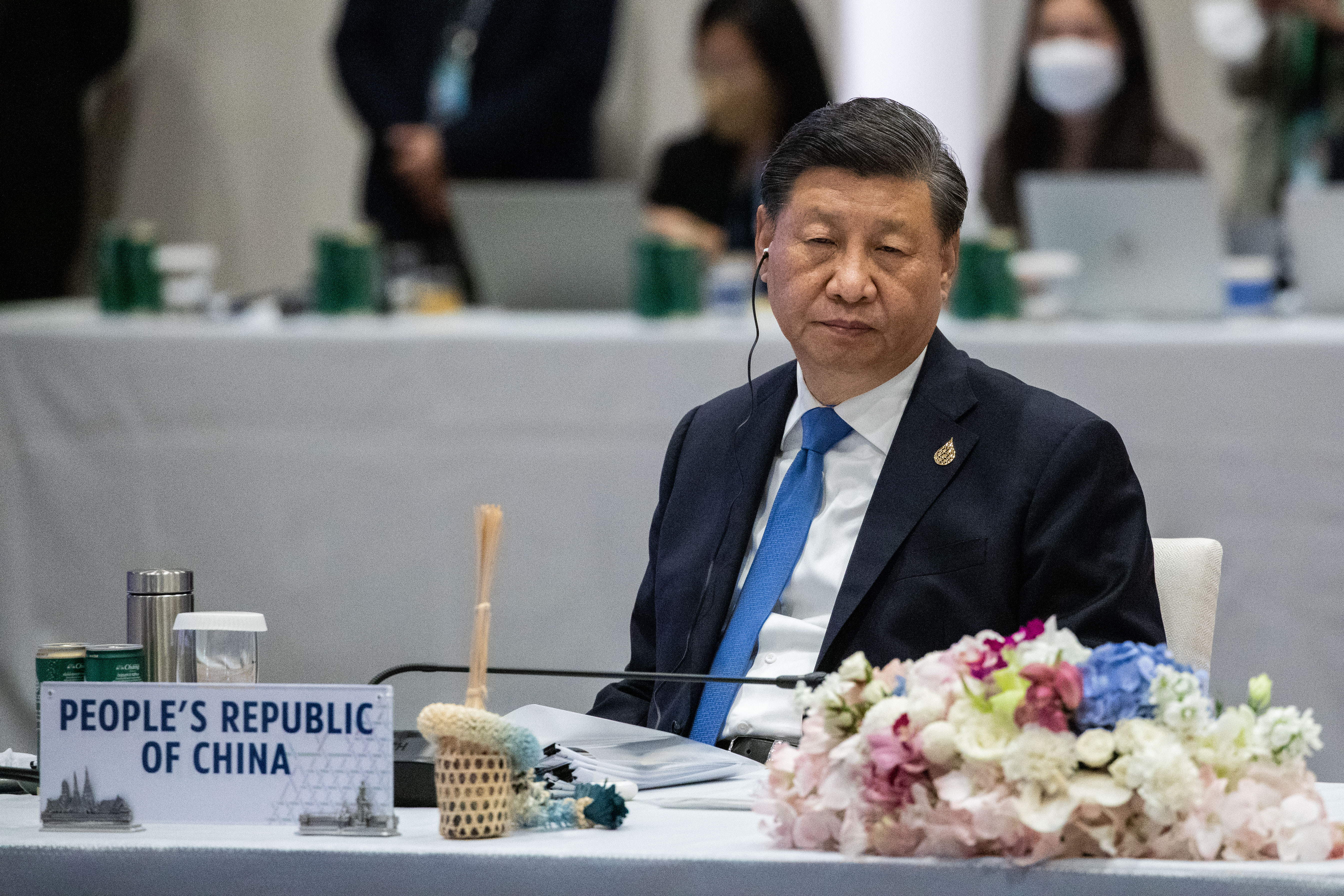 Xi Jinping sits at a table. A placard reads “People’s Republic of China” on the table in front of him. On his face is an expression of boredom, or perhaps weary tiredness. 