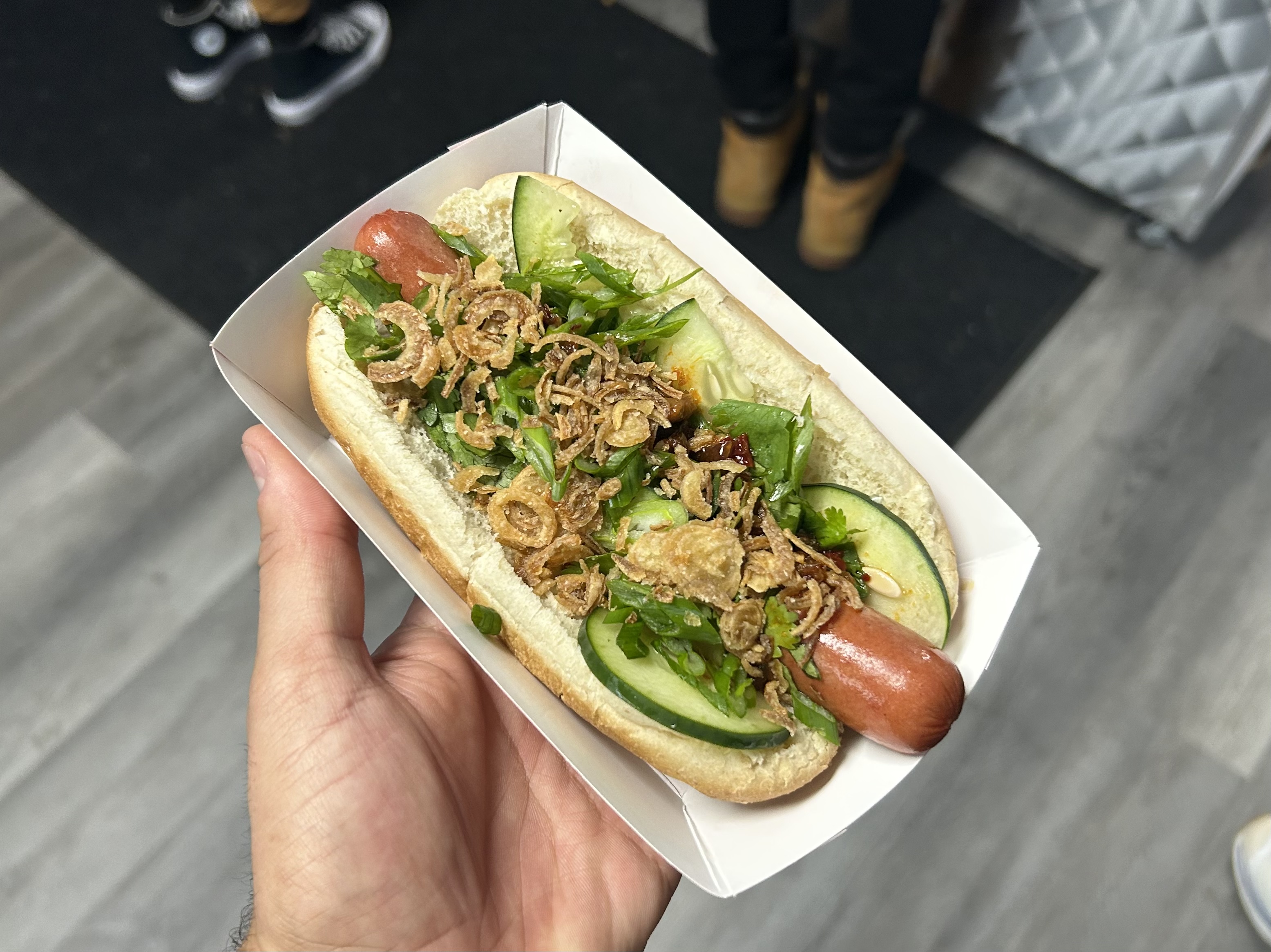 A hot dog loaded with fried shallots, chili crisp, cilantro, and cucumber.