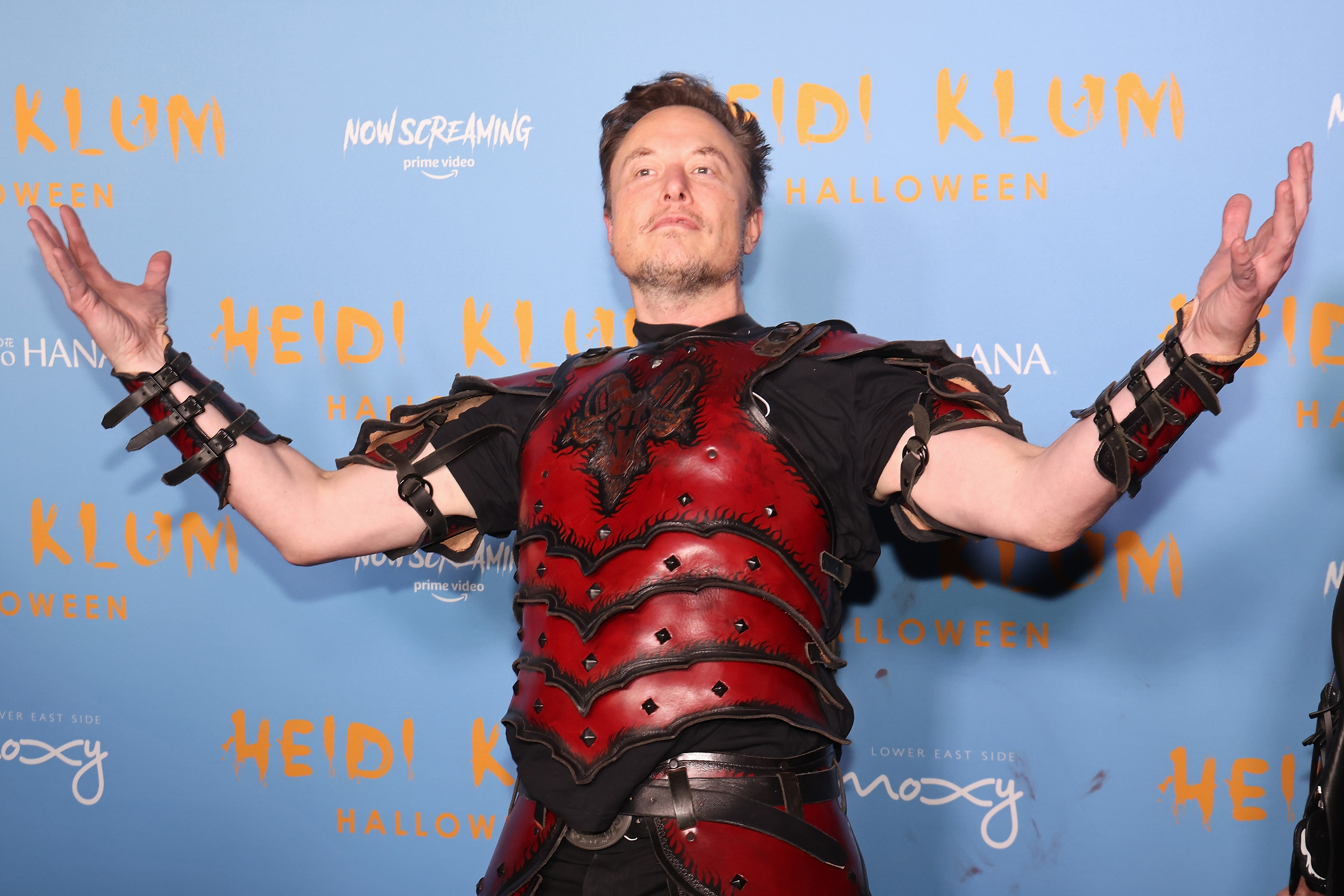 Elon Musk dressed in a gladiator-style costume, holding his arms up and out in a “look at me” gesture.