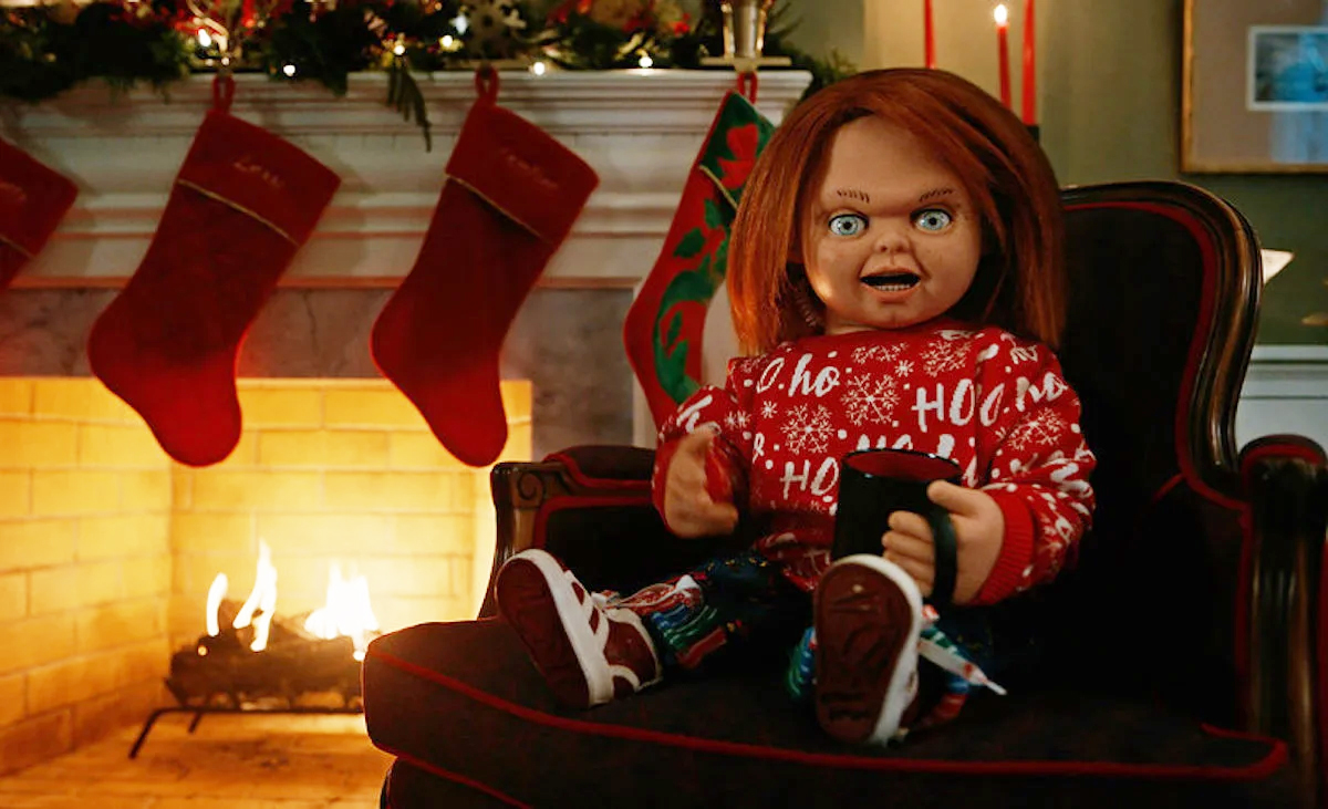 Chucky the doll wears a Christmas sweater by the fire place with a mug on his lap.
