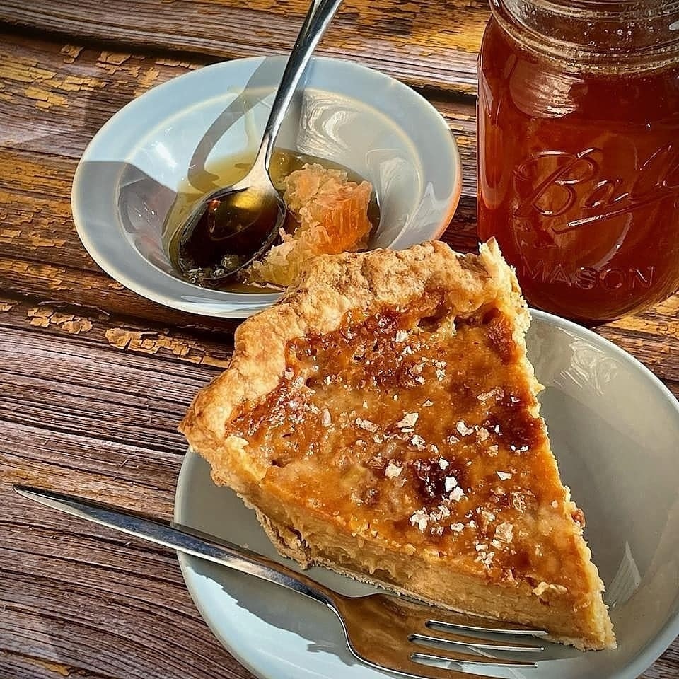A slice of honey pie on a plate in front of a mason jar of honey and small dish containing honey and honeycomb.