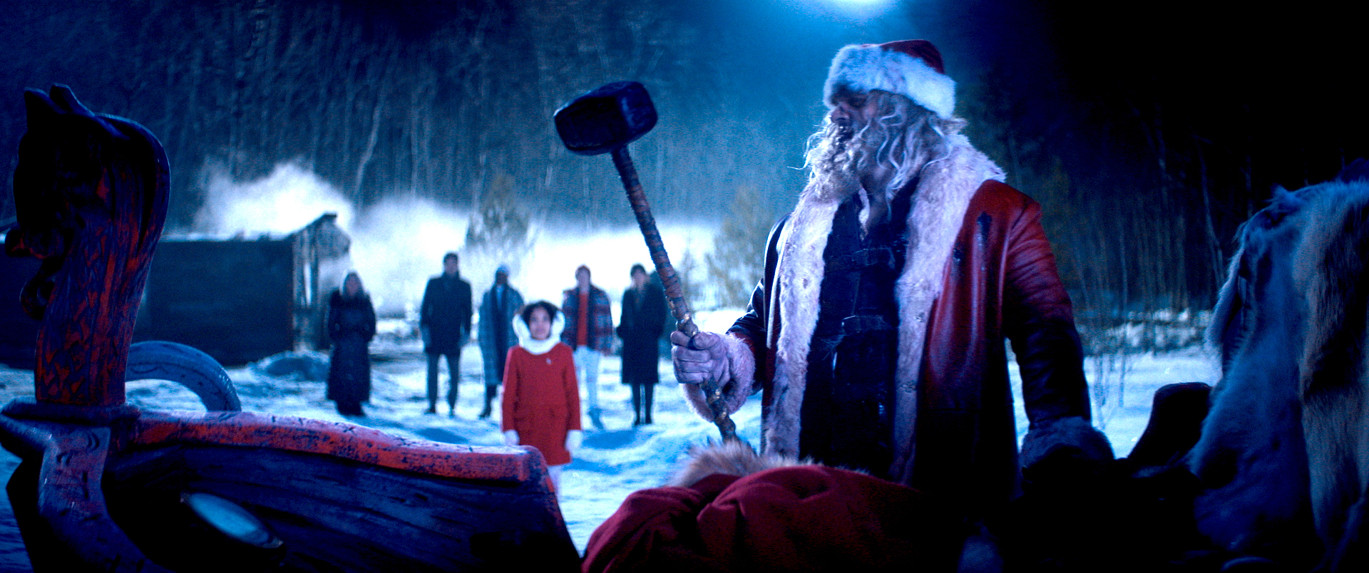 Santa Claus (David Harbour) stands in front of the silhouette of his sleigh outdoors at night, holding up a sledgehammer as a crowd looks on in Violent Night