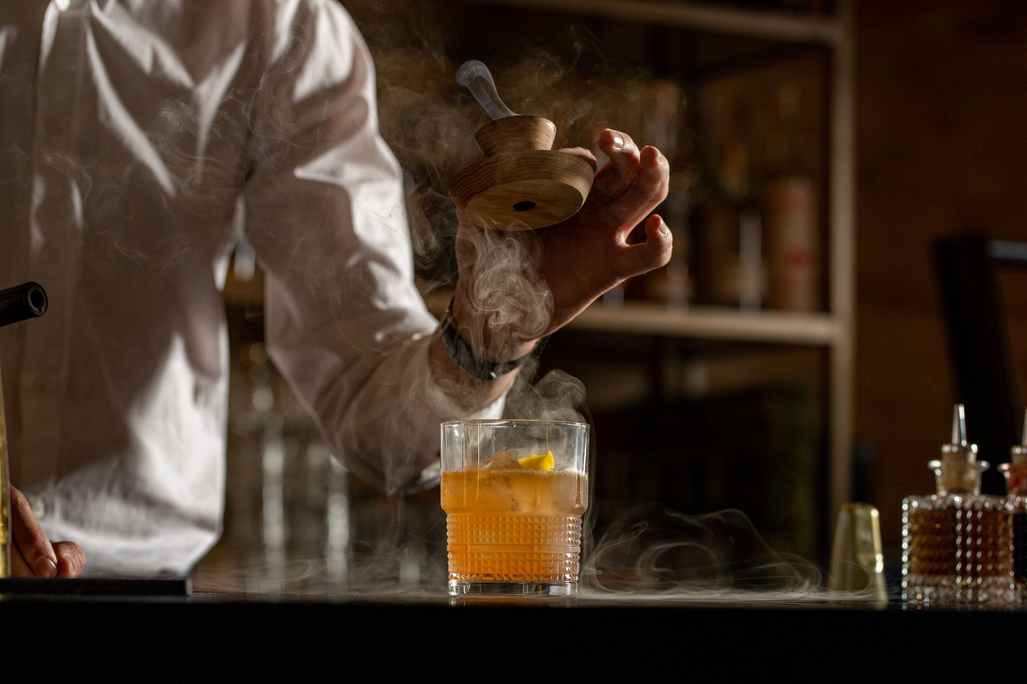 A hand lifts a wooden stopper from on top of a cocktail glass at a dim bar.