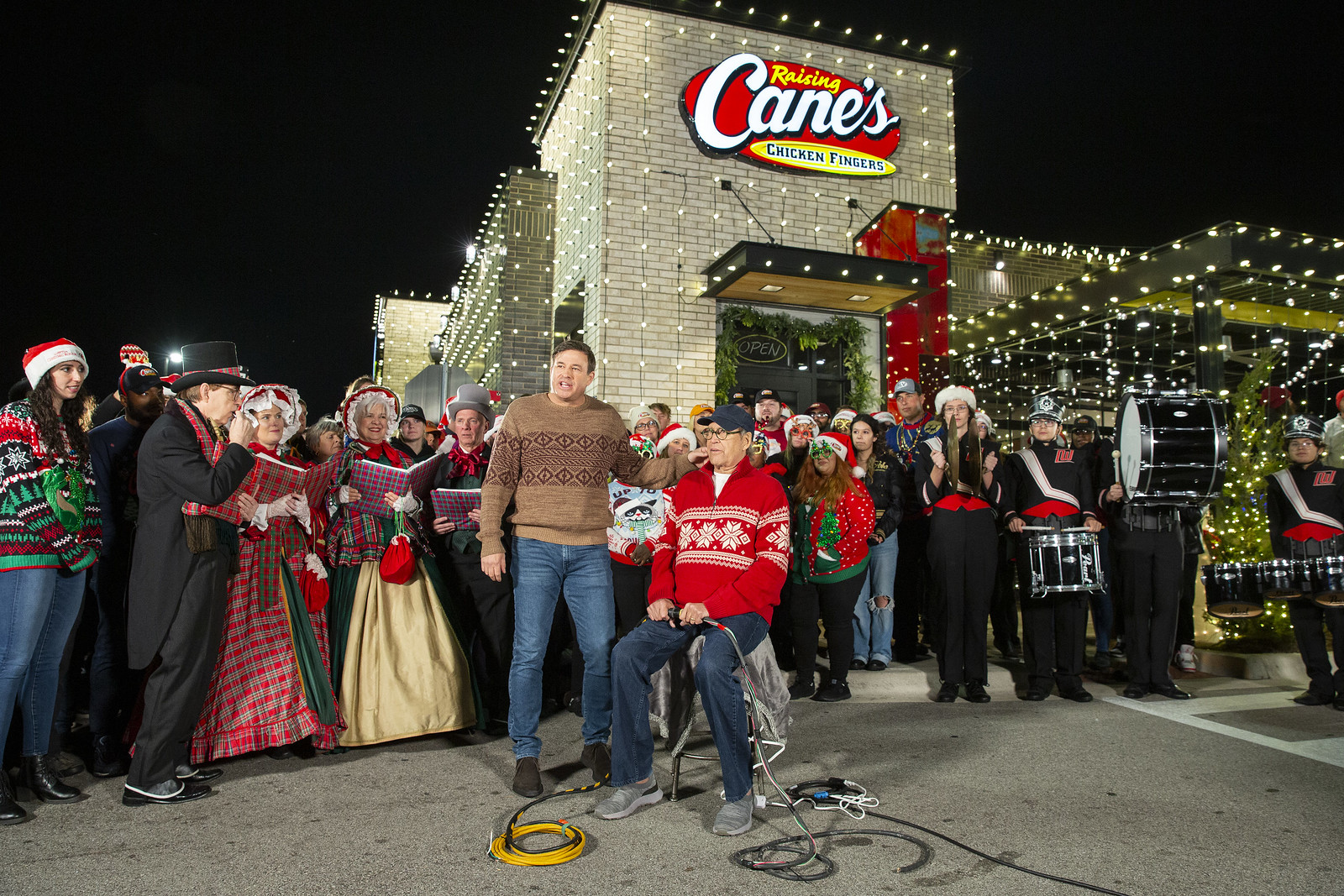 Two men in sweaters perform a scene in front of a group of people dressed in Christmas Carol outfits. They are outside of a Raising Cane’s restaurant.