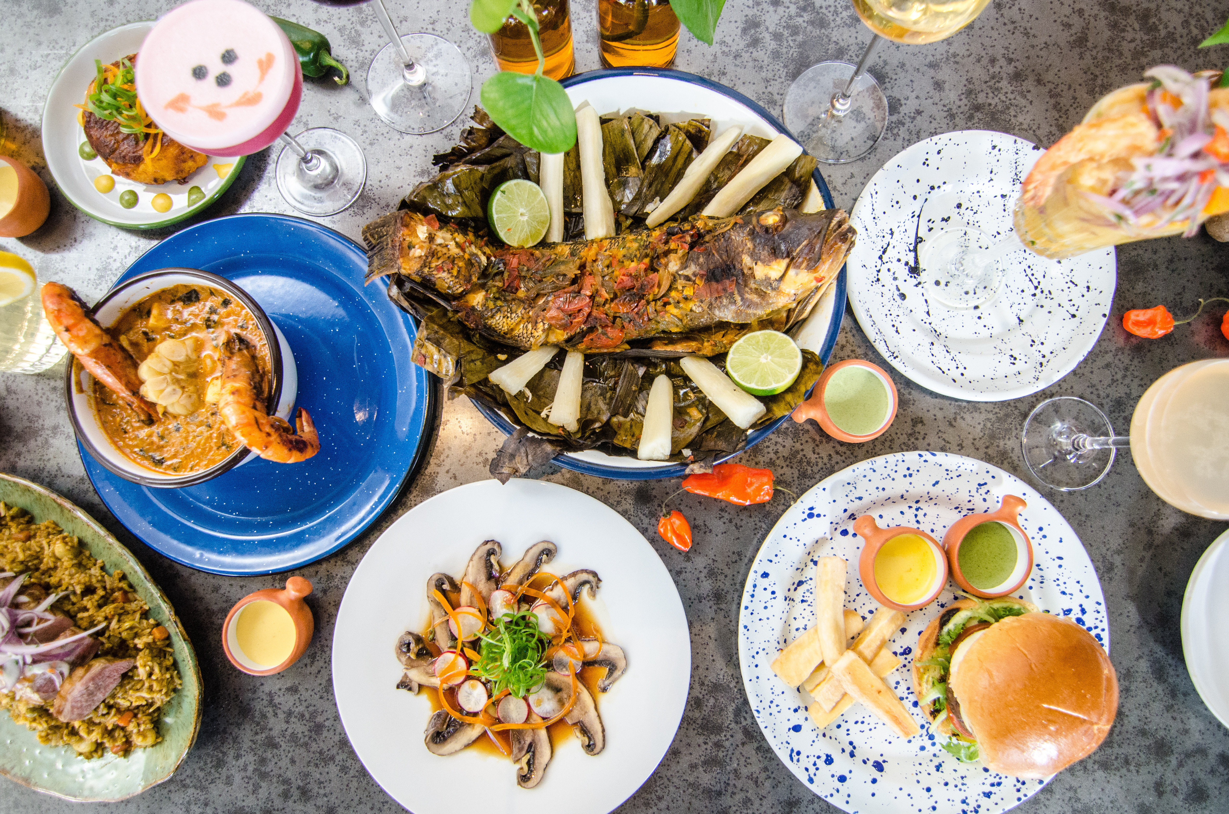Overhead view of an array of colorful dishes on a metallic table surface, including a whole grilled and steamed fish, a burger, two giant prawns coming out of a bisque, and more.