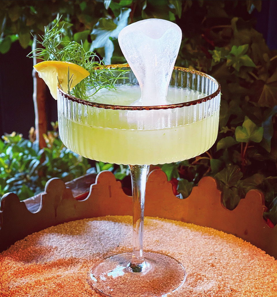 A lime green cocktail in a fancy coupe class with an ice cube in the shape of a cobra. The glass is placed on a bed of sand.