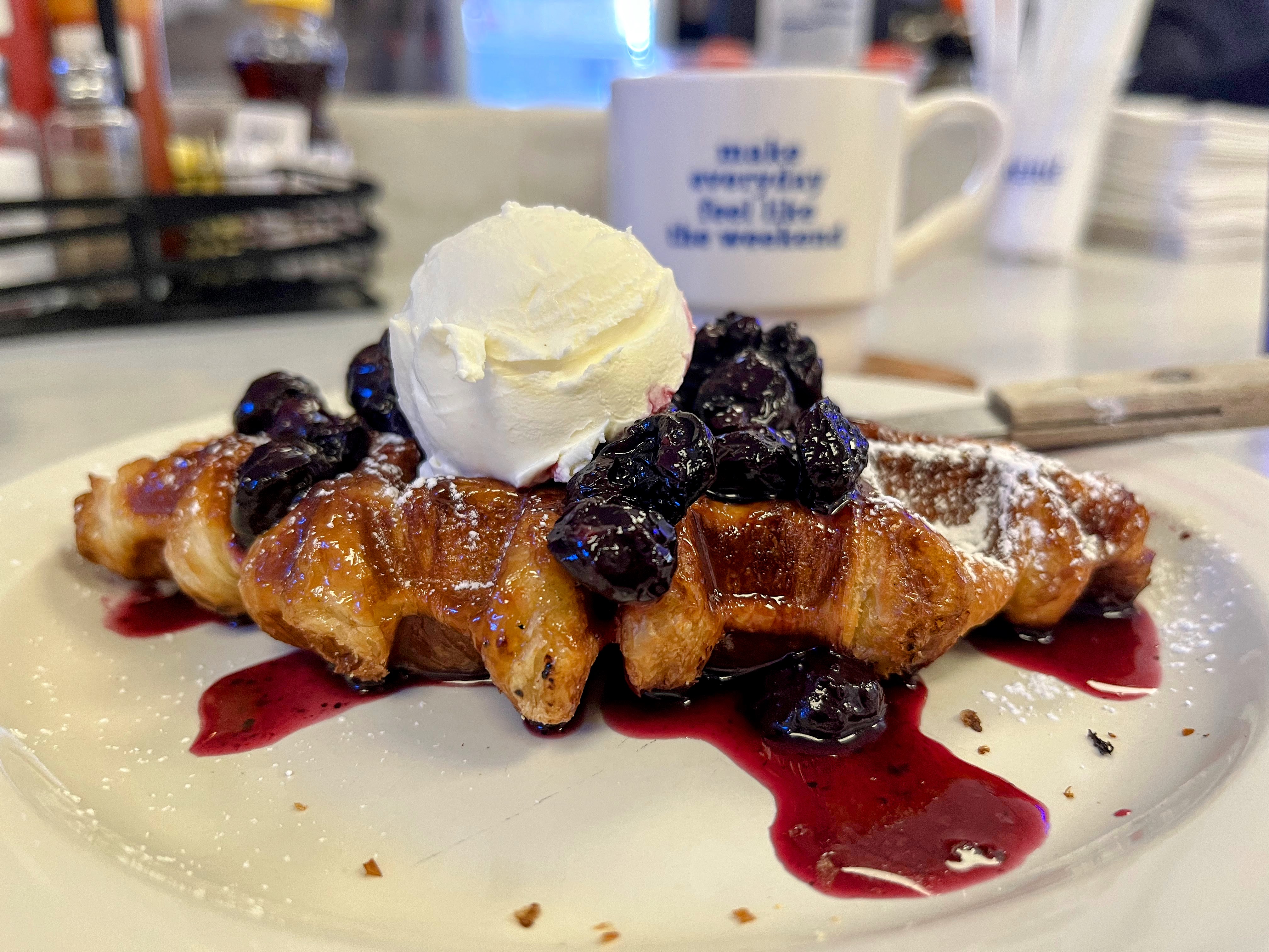 A croissant pressed into a waffle shape is drizzled in blueberry jam and a scoop of whipped cream.