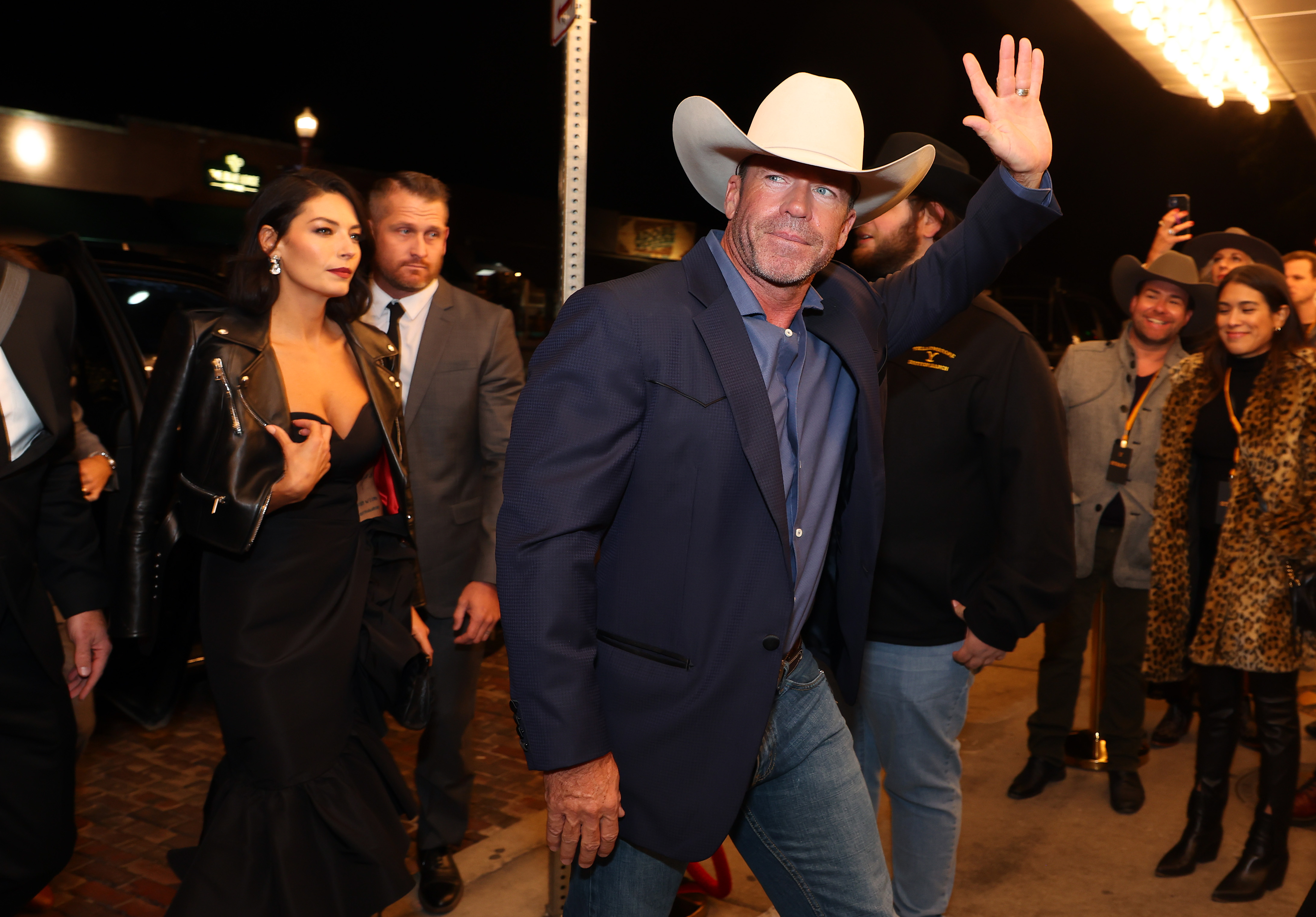 Taylor Sheridan wears a white cowboy hat, jeans, blue blazer, and light blue button up shirt. His wife Nicole, wears a black strapless full-length gown with a leather jacket.