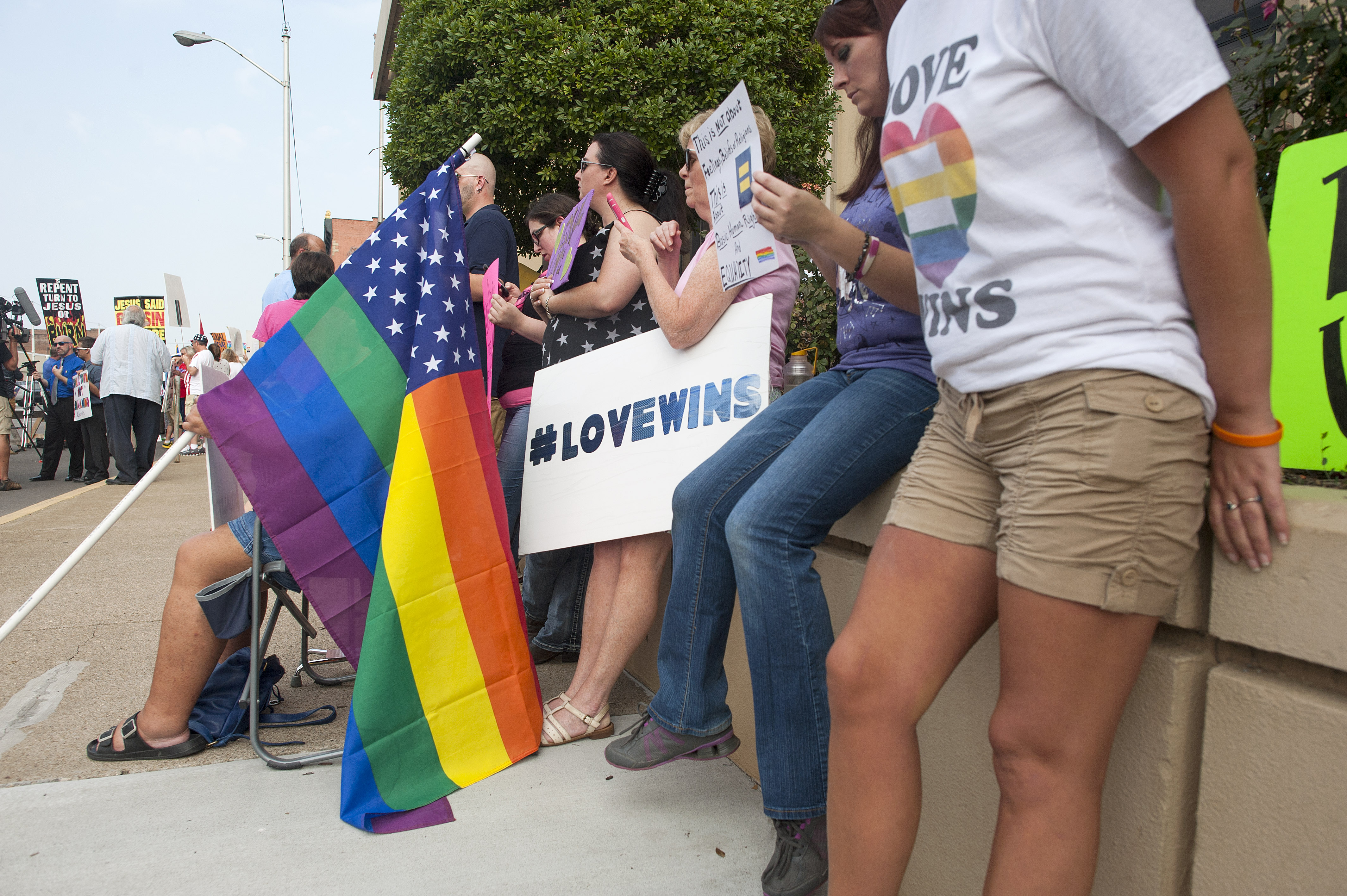 People gather on a tree-lined street, standing and sitting in lawn chairs, wearing shirts and holding signs that support same-sex marriage rights, including “Love wins” and Pride symbols. 