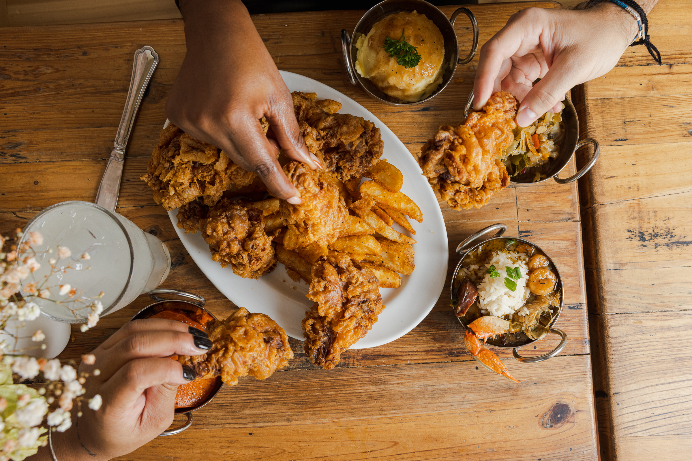 Hands reach in for fried chicken on a wide plate atop a wooden table.