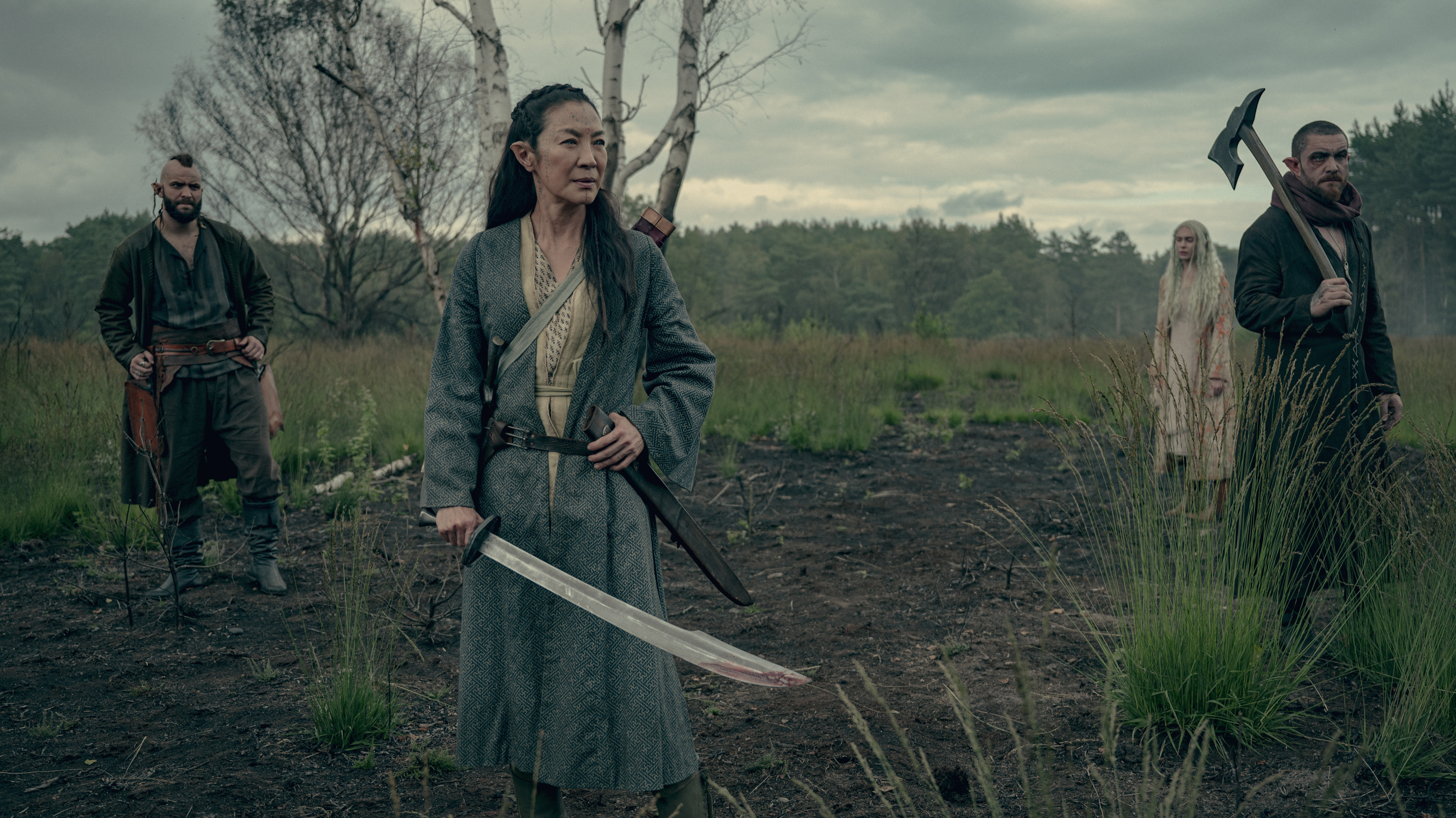 Scian (Michelle Yeoh) standing with her sword; in the background is Brother Death (Huw Novelli), Zacare (Lizzie Annis) and Fjall (Laurence O’Fuarain)