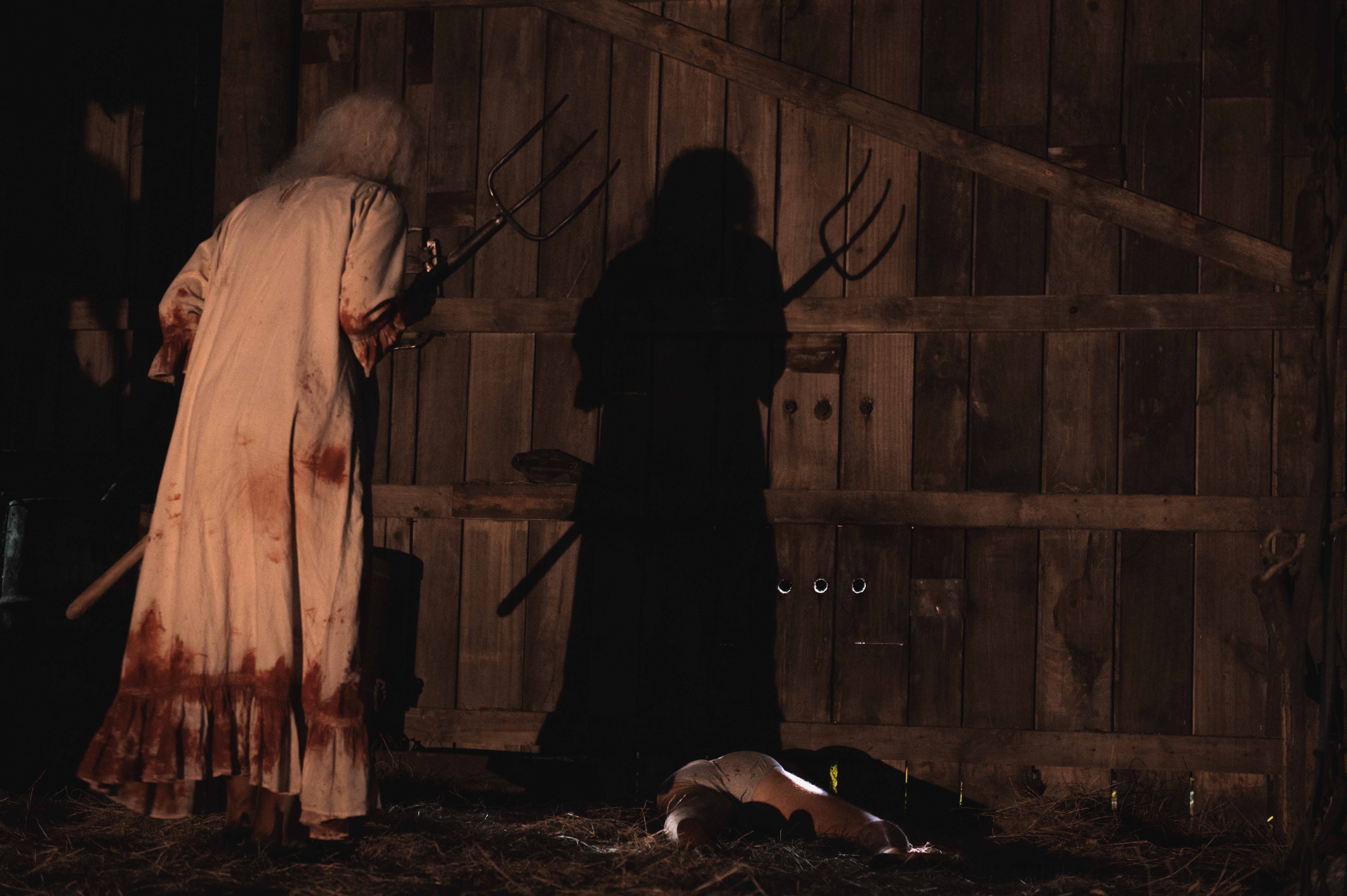 A dark-lit, creepy scene shows an old woman with long white hair in a long white dress with bloodstains covering it, hunched over and holding a pitchfork, her shadow cast on a wooden barn wall in front of her.