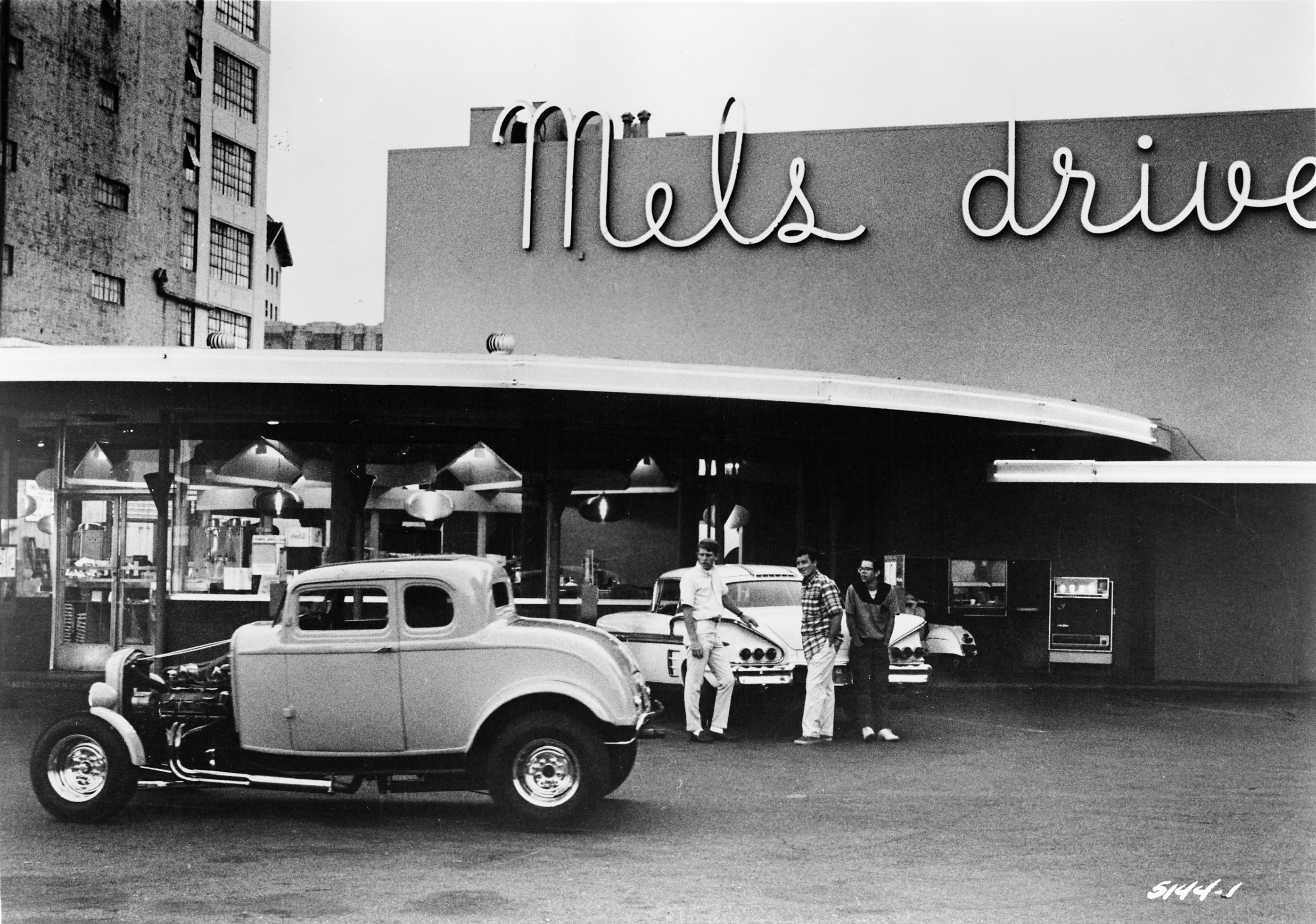 A black and white vintage photograph of a hotrod car with open hood in front of a diner.
