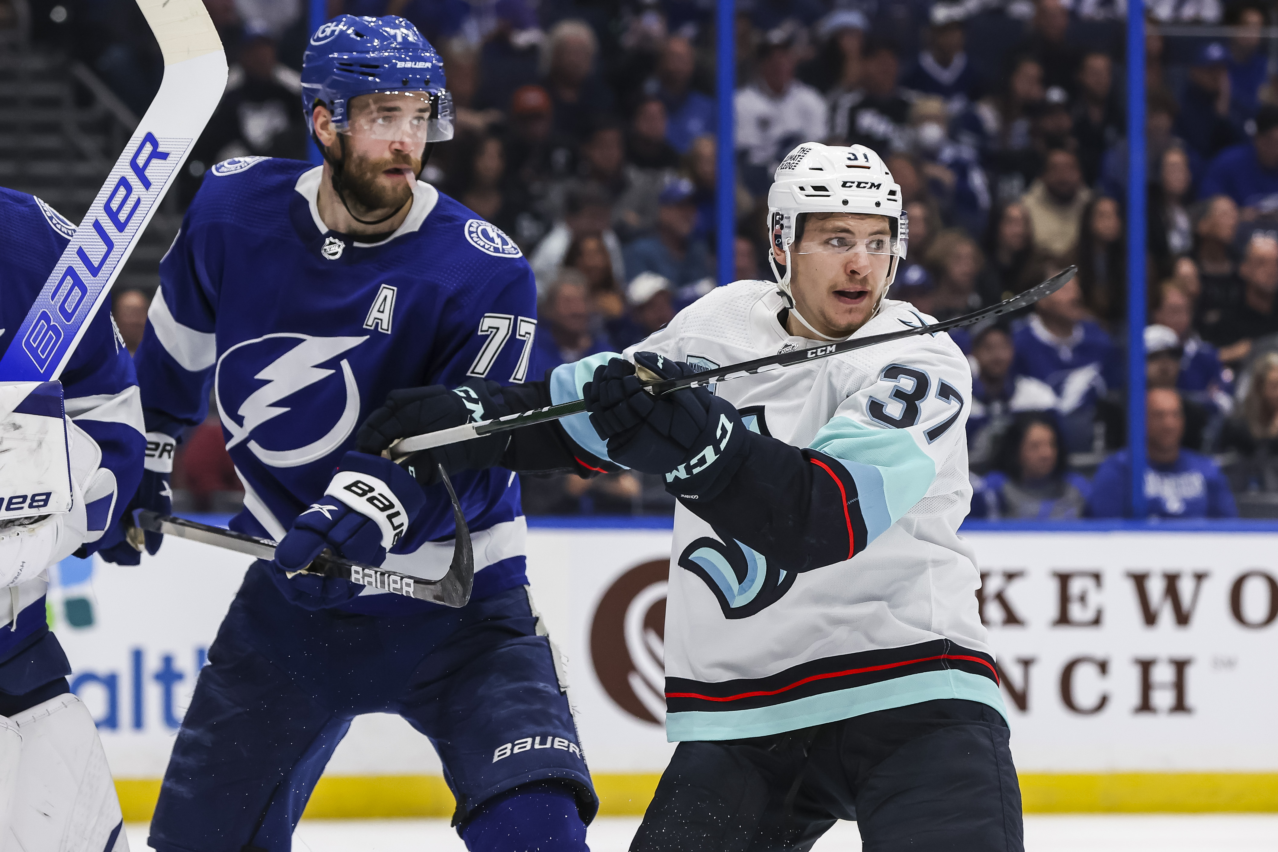 A mid-range shot of Victor Hedman (left) in a blue home jersey and Yanni Gourde (right) in a white away jersey. Both players are looking towards the right-hand frame of the shot.