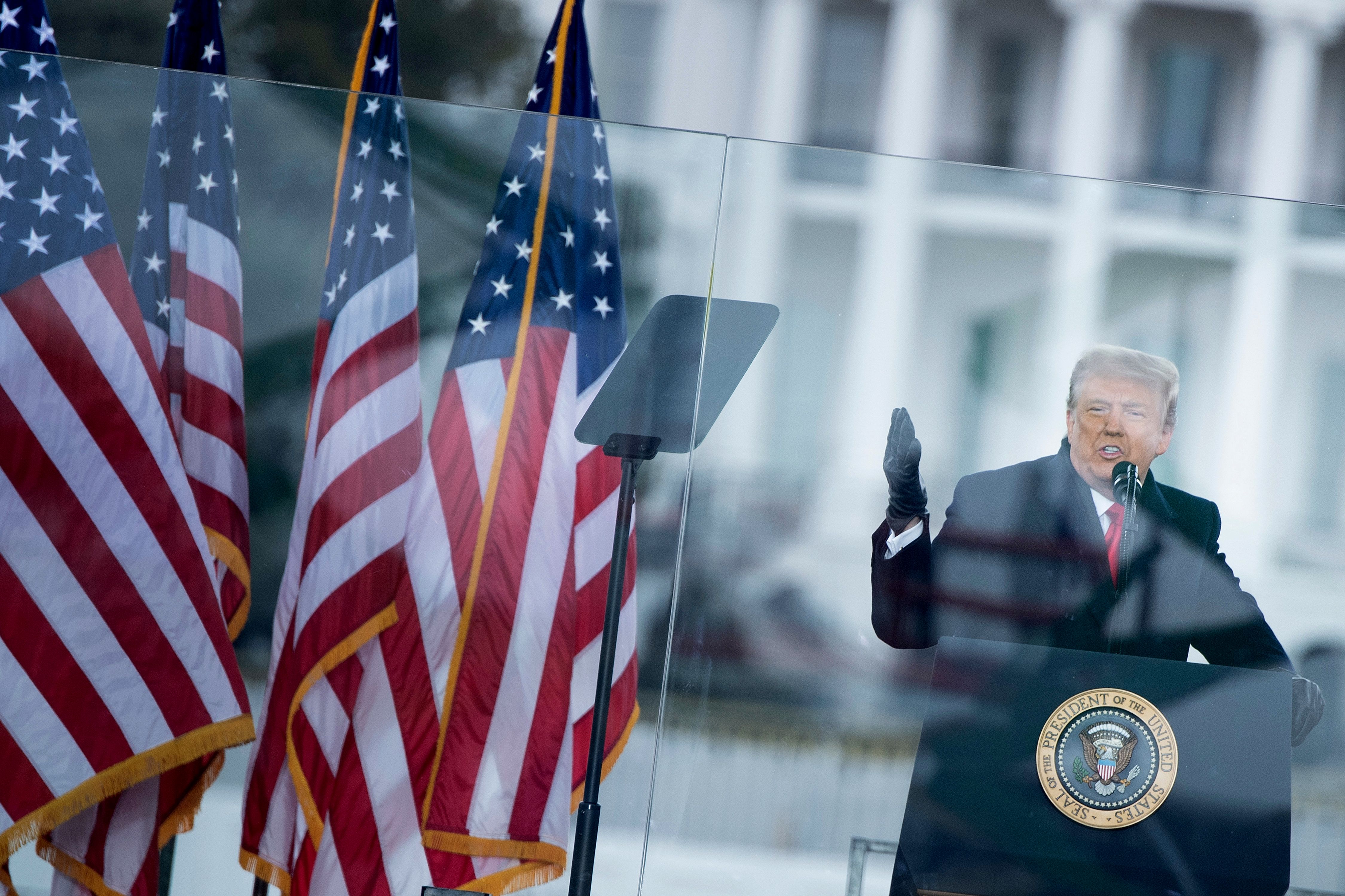 Trump, in a black overcoat and black leather gloves, speaks behind bulletproof glass, a row of US flags behind him. The camera is tilted, making it seem as if Trump is about to tilt over.